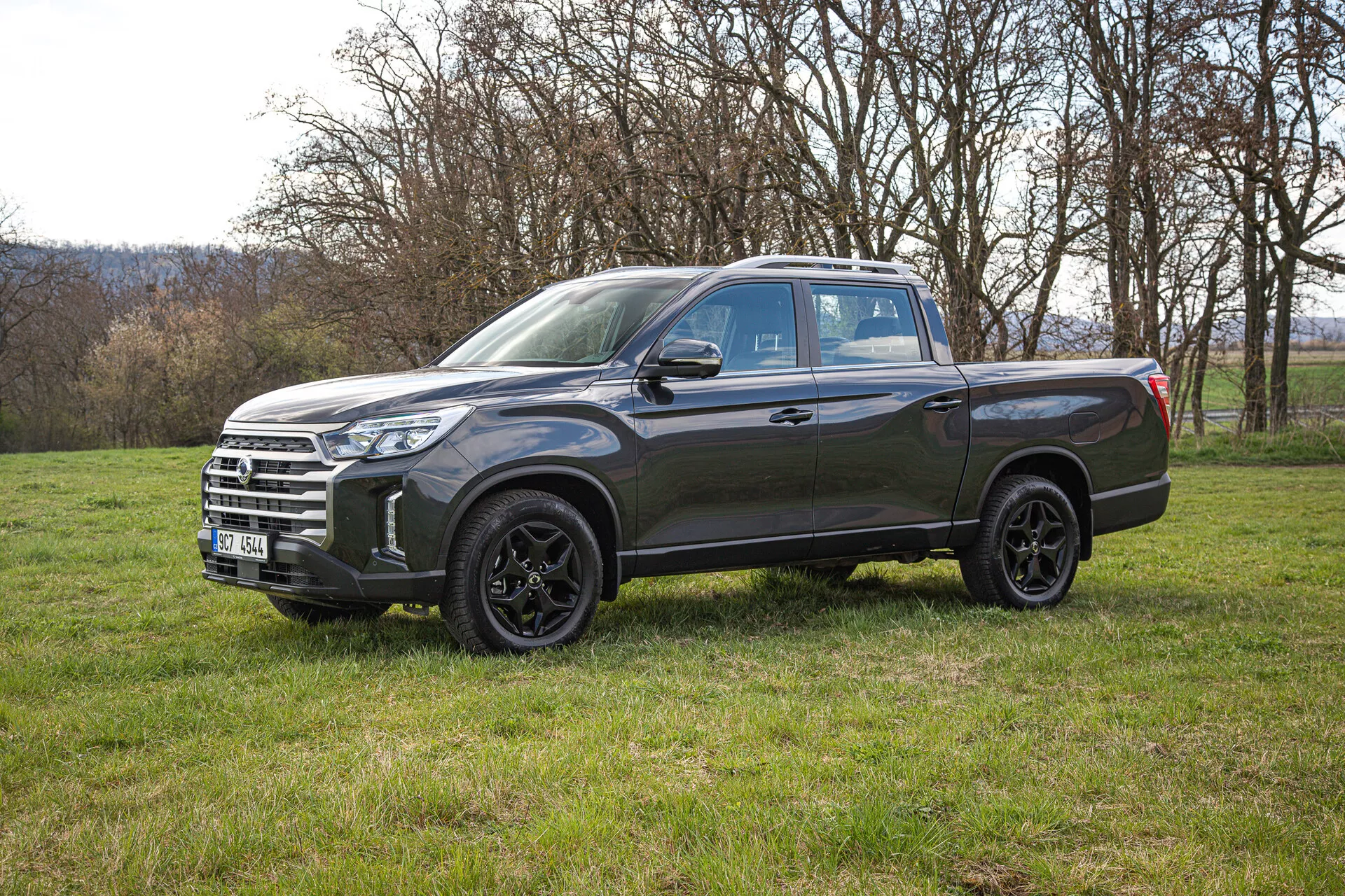 Vue latérale d'un pick-up SsangYong Musso Grand 2022 de couleur gris foncé sur un terrain herbeux, mettant en avant ses roues en alliage, phares LED et grand espace de chargement