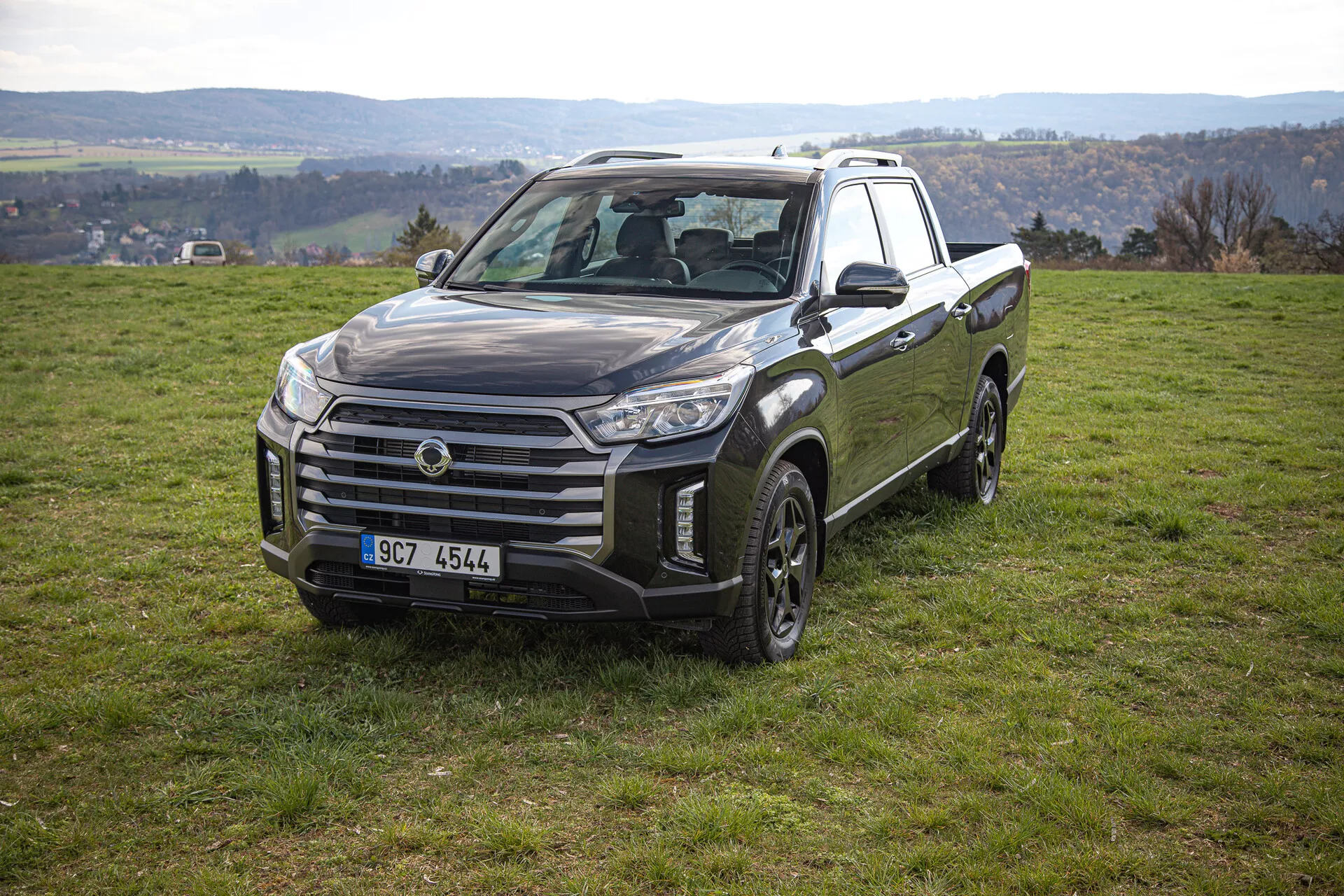 Pickup SsangYong Musso Grand 2022 en position stationnaire sur une pente herbeuse montrant la vue de face et latérale incluant une grande grille, des blocs optiques angulaires, et un design de roue apte au tout-terrain, avec un paysage rural en arrière-plan.
