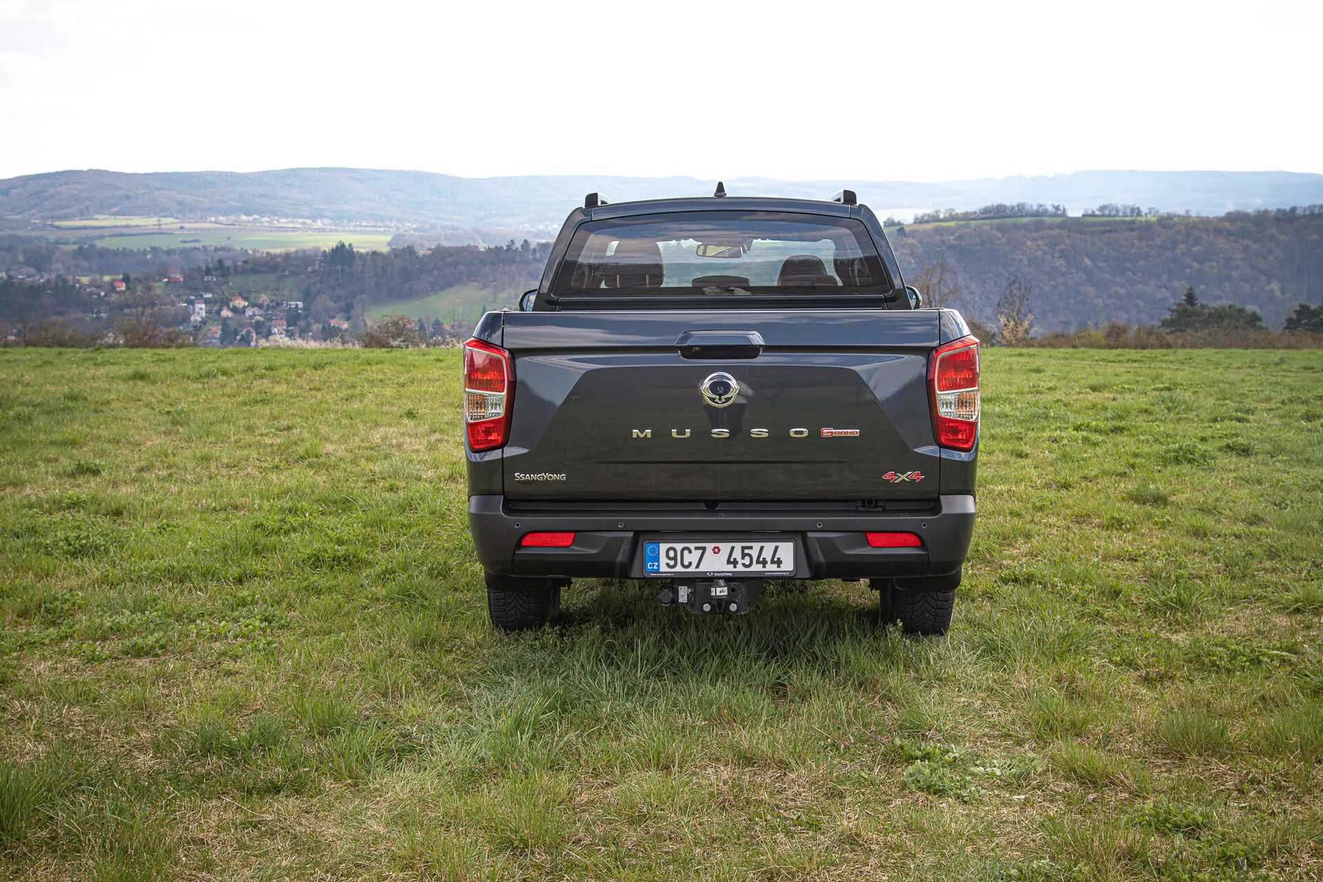 Vue arrière d'un pick-up SsangYong Musso Grand 2022 de couleur foncée sur un terrain herbeux inégal avec un paysage rural en arrière-plan, montrant la marque sur le hayon, les feux arrière verticaux, les capteurs arrière et l'attelage de remorque en journée