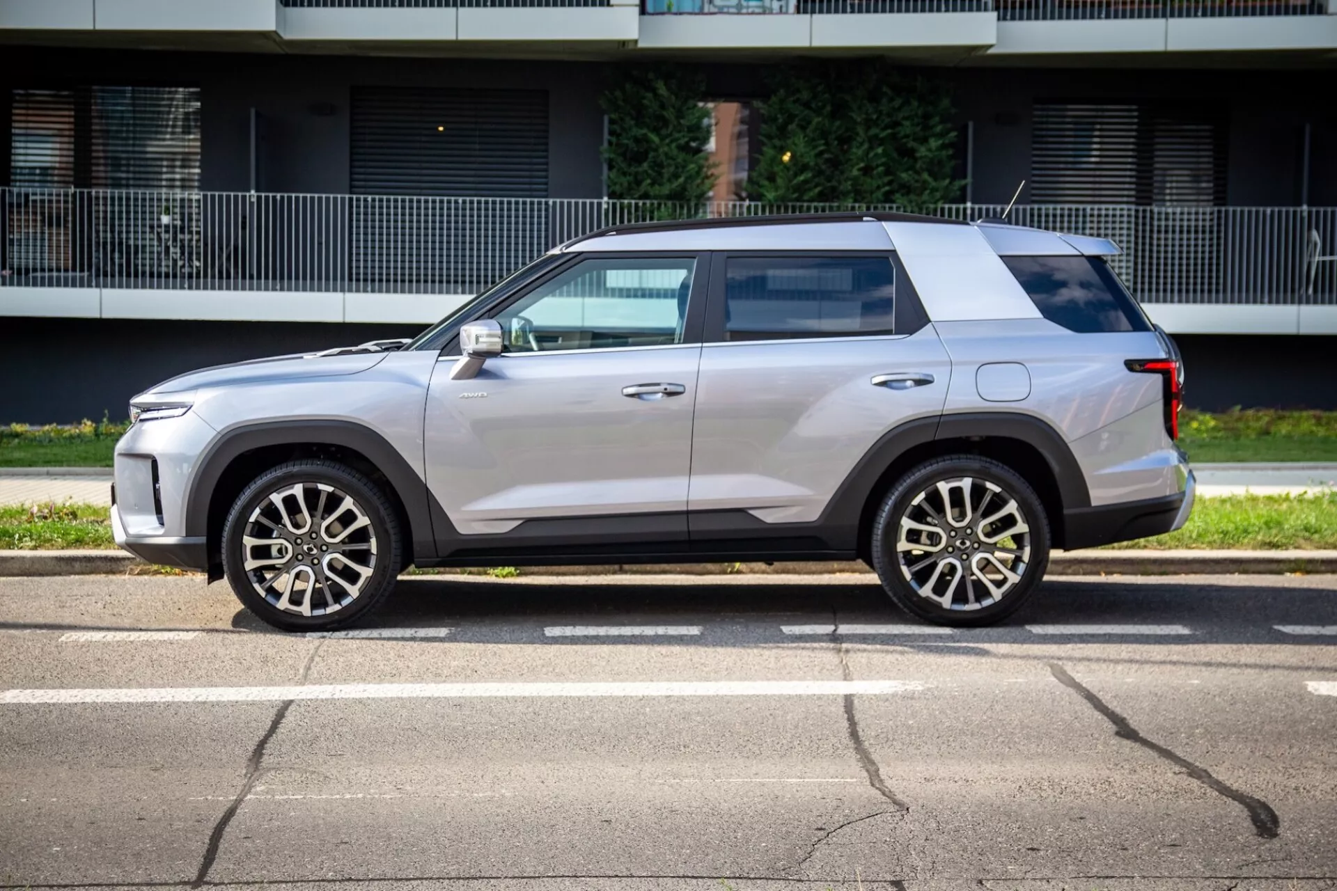 Vue de profil du SsangYong Torres 2023 montrant sa conception carrée mais aérodynamique, ligne de ceinture haute, toit plat et peinture métallique claire avec jantes en alliage à cinq branches, rétroviseurs latéraux couleur carrosserie avec clignotants intégrés, légère teinte des fenêtres, poignées de porte affleurantes et garniture noire dans une rue ensoleillée