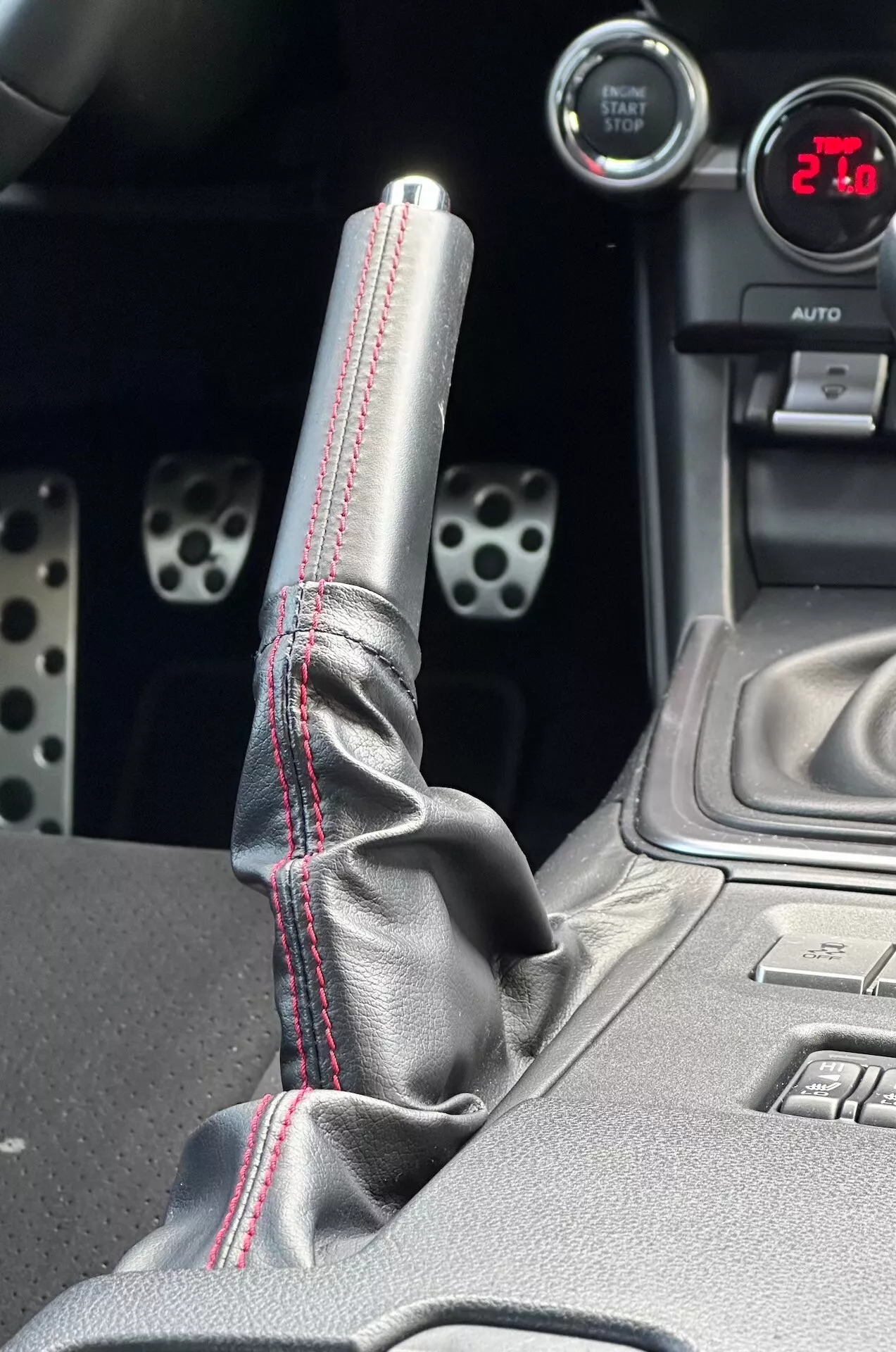 Vue rapprochée de la console centrale de la Subaru BRZ 2023 mettant en avant le levier de vitesse en métal avec coutures rouges contrastantes sur le soufflet en cuir, jauge numérique affichant '21', pédales sportives en aluminium avec des trous percés et des fonctionnalités technologiques telles que boutons de contrôle de la stabilité électronique.