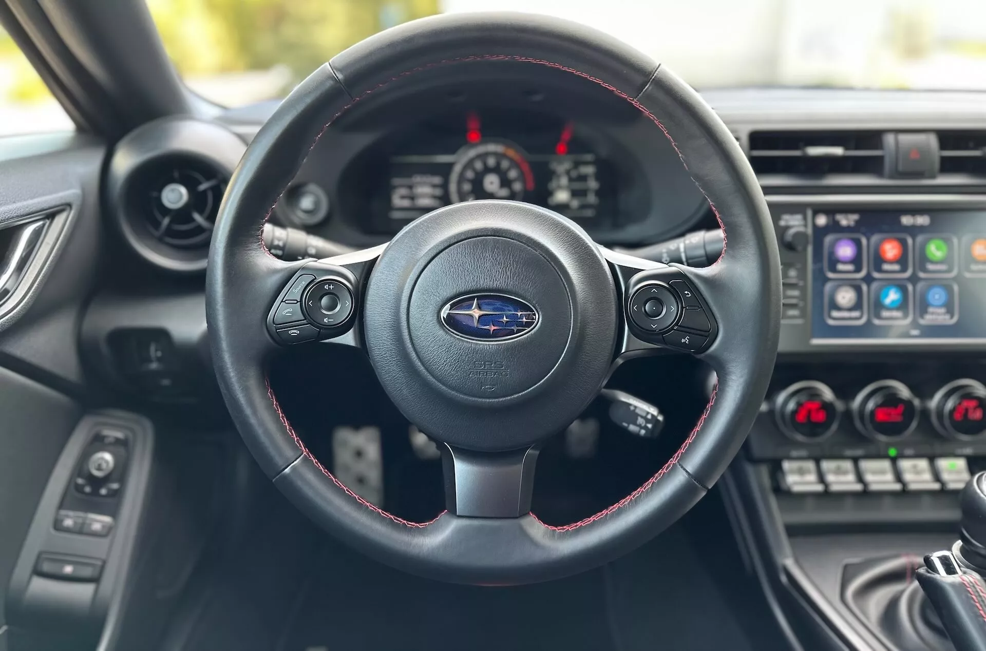 Vue intérieure du Subaru BRZ 2023 depuis le siège du conducteur se concentrant sur le volant à trois branches avec coutures rouges, logo Subaru, et tableau de bord avec cluster d'instruments et console centrale