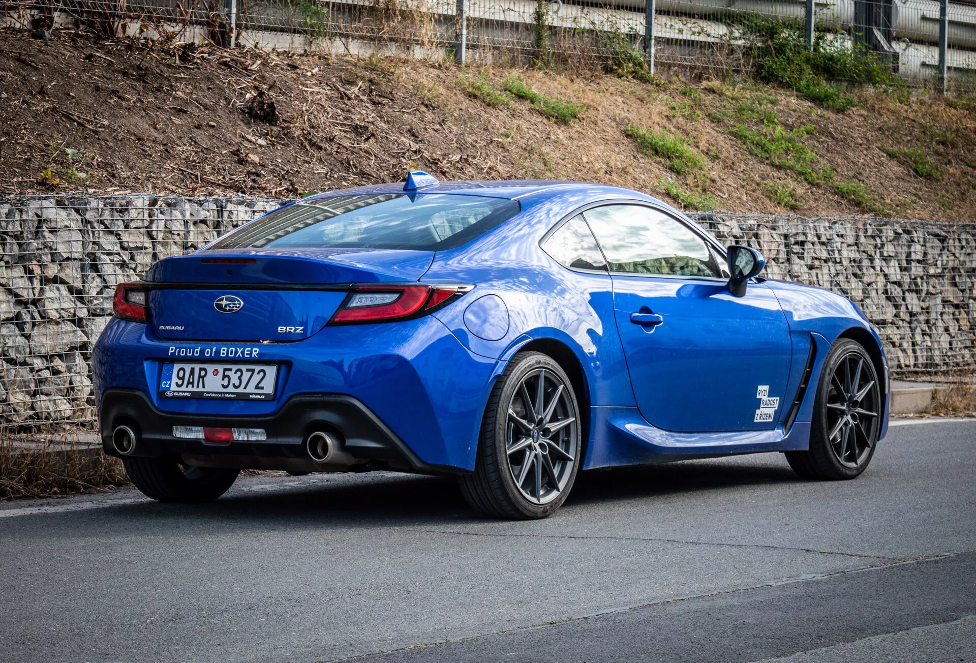 Coupé sportif Subaru BRZ 2023 en bleu métallique garé sur une pente, présentant sa vue arrière trois quarts mettant en évidence le profil latéral du véhicule, l'arrière courbé, les roues en alliage sportives, les embouts d'échappement doubles, le petit spoiler et le badge du moteur 'Proud of Boxer'.