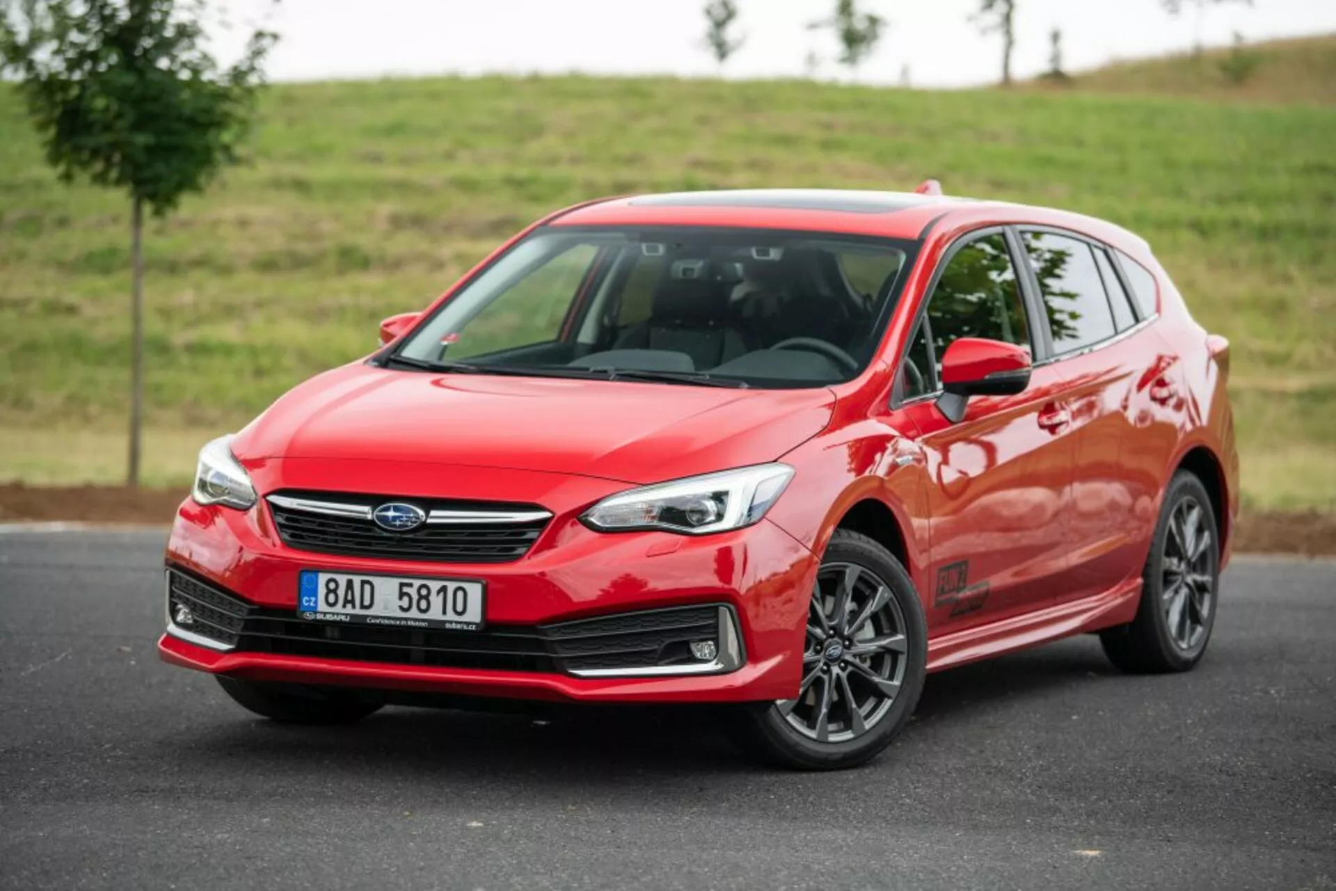 Subaru Impreza hatchback rouge vif de 2018 avec vue de trois quarts avant, présentant ses phares LED élégants, sa grille distinctive avec emblème central, et ses jantes en alliage à multiples rayons contre un arrière-plan naturel nuageux