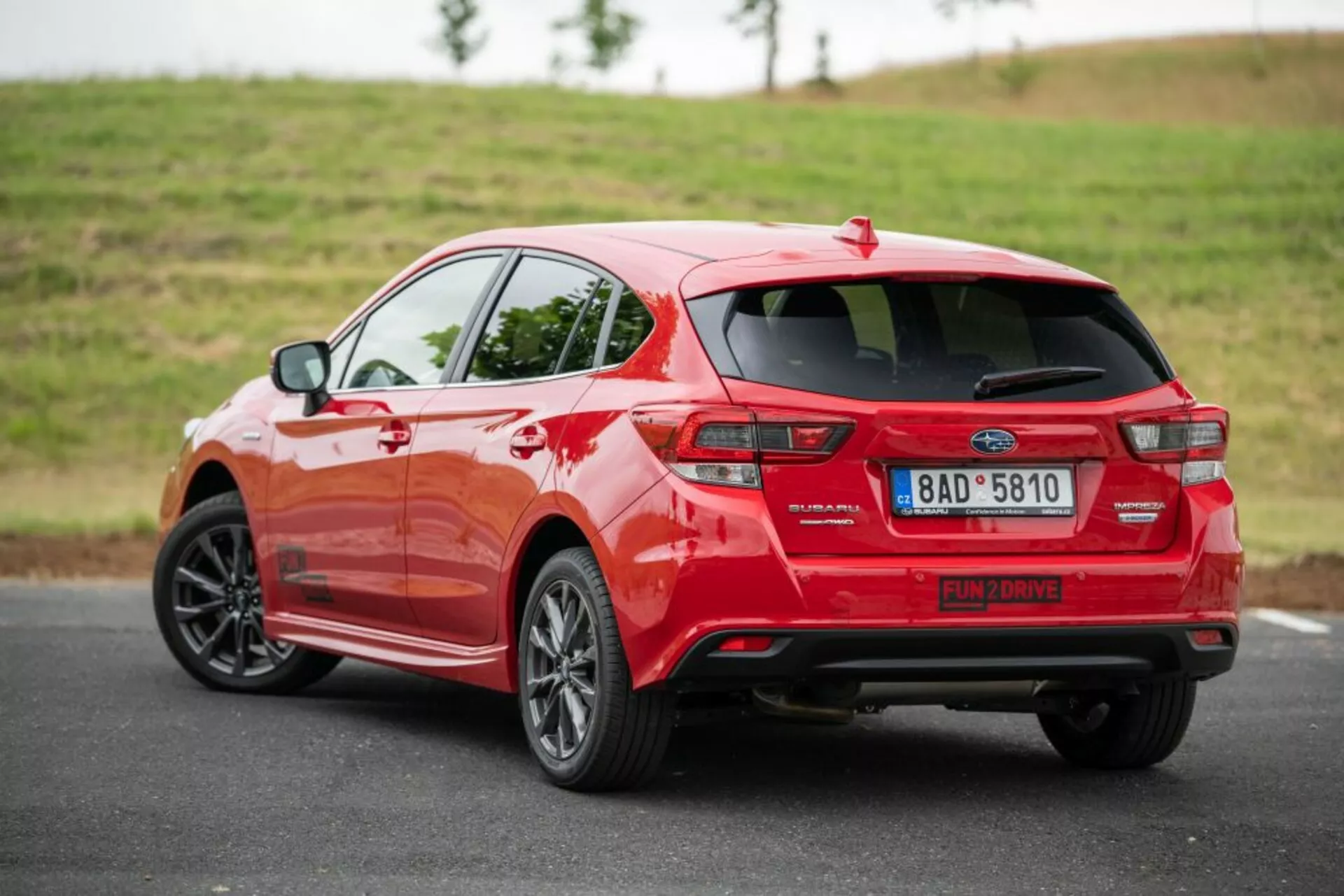 Hatchback rouge Subaru Impreza 2018 vu depuis un angle arrière trois-quarts mettant en évidence son profil latéral, le design distinctif des feux arrière, le système d'échappement, le spoiler et les jantes en alliage sur une route en asphalte avec un arrière-plan gazonné dans une lumière douce et couverte