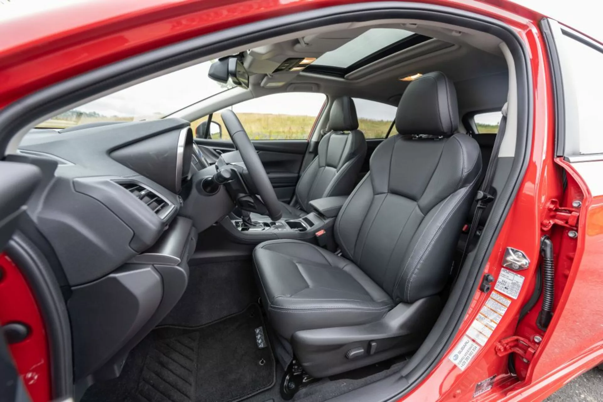 Vue intérieure de la Subaru Impreza 2018 depuis une perspective extérieure montrant le siège conducteur, le tableau de bord, le volant avec boutons de contrôle, sièges avant ergonomiques en cuir noir et commandes intérieures de porte à travers la porte du conducteur ouverte