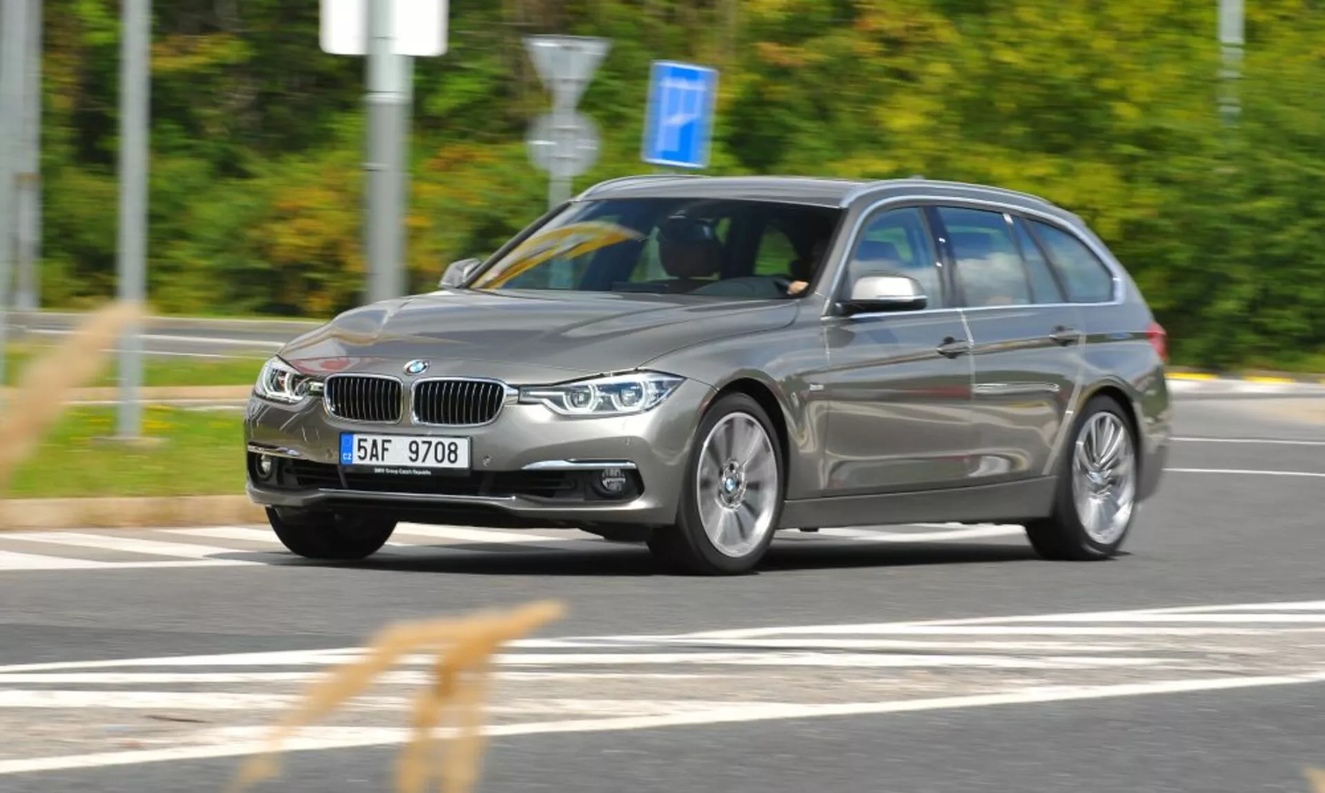 Vue dynamique trois quarts avant d'un break BMW argenté en mouvement avec grille réniforme distinctive et emblème, capturée d'un angle légèrement élevé indiquant vitesse et mouvement, se concentrant sur les caractéristiques de design et technologie comme phares angulaires, roues en alliage et barres de toit dans un cadre extérieur flou de mouvement.