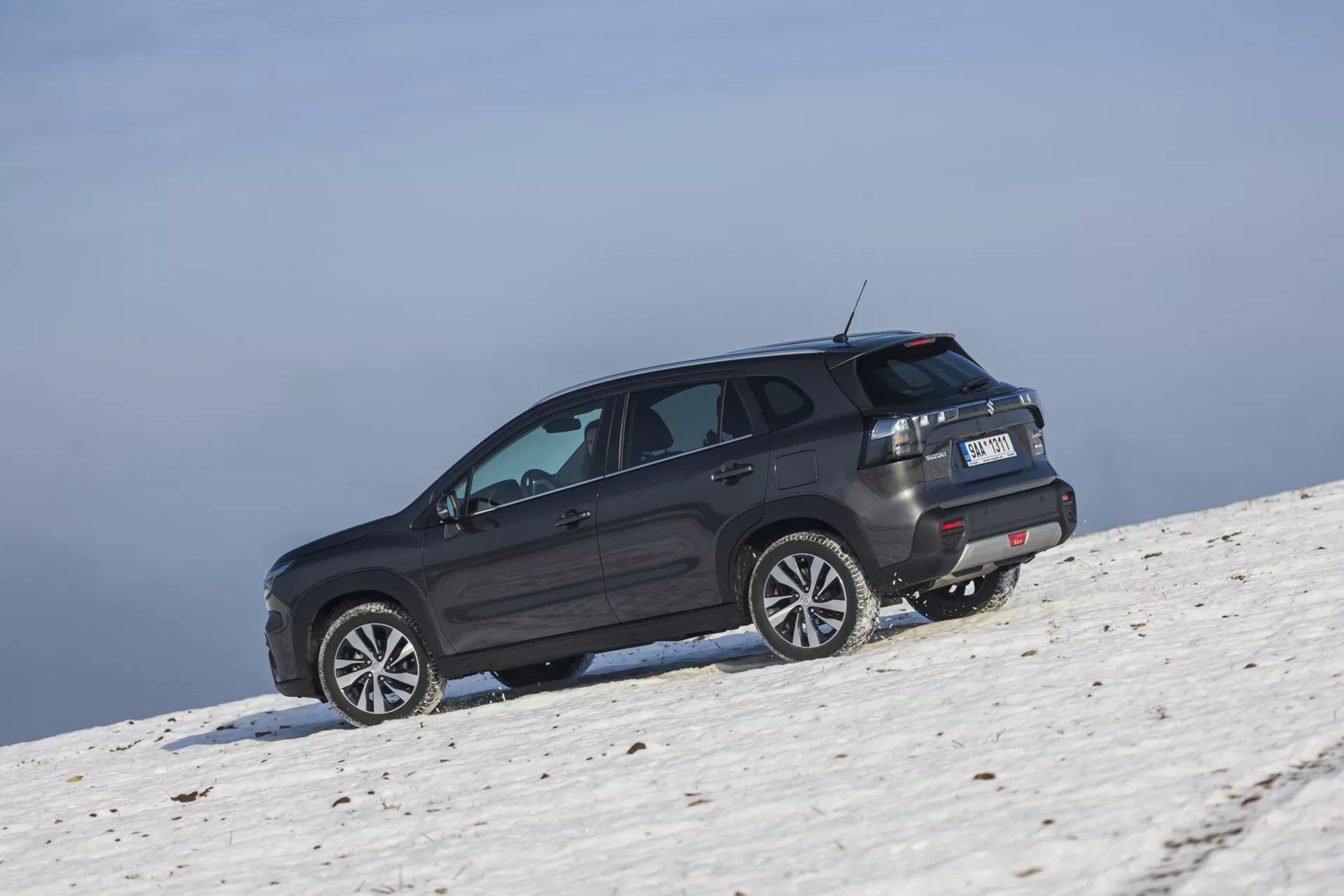 SUV Suzuki S-Cross 2022 de teinte sombre positionné sur une pente enneigée capturé depuis un angle arrière oblique soulignant son profil latéral et arrière, grandes roues en alliage, vitres teintées avec dégivreur, feux arrière LED, aileron de toit, et des capacités potentielles tout-terrain sous des conditions de ciel clair.