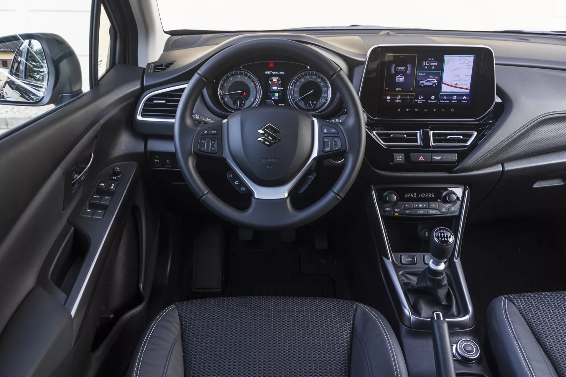 Intérieur de la cabine avant du Suzuki S-Cross 2022 montrant le tableau de bord, le volant avec commandes intégrées, le tableau de bord numérique-analogique, le système d'infodivertissement tactile, les commandes du climatiseur, le levier de vitesse en cuir, le sélecteur de mode de conduite, le frein de stationnement électronique et le panneau de porte avec boutons de commande