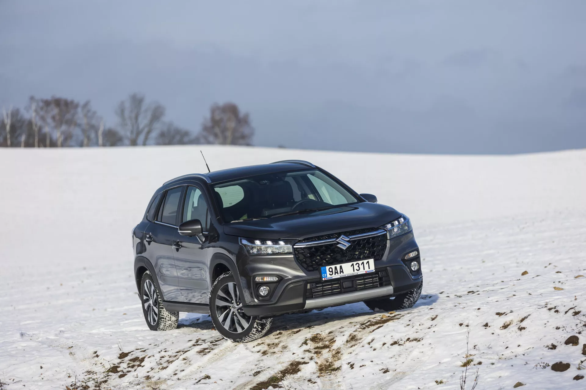 SUV compact Suzuki S-Cross de couleur sombre, modèle 2022, garé dans un paysage enneigé, présentant une vue de face et de côté à un angle de 45 degrés, mettant en avant une grille élégante avec finition métallique, phares LED allongés, jantes en alliage à multiples branches, et un encadrement de fenêtre chromé poli indiquant un pack luxe et une capacité potentielle tout-terrain