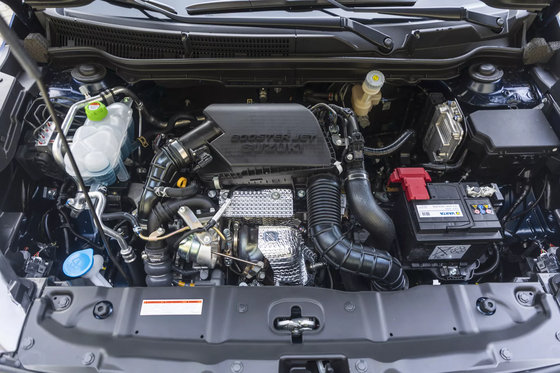 Compartiment moteur de la Suzuki S-Cross 2022 présentant une vue de face avec des composants visibles tels que le bloc moteur, la culasse, le collecteur d'admission et des caractéristiques technologiques incluant le turbocompresseur et le calage variable des soupapes, ainsi que le branding Suzuki sur le couvercle du moteur, en état impeccable.