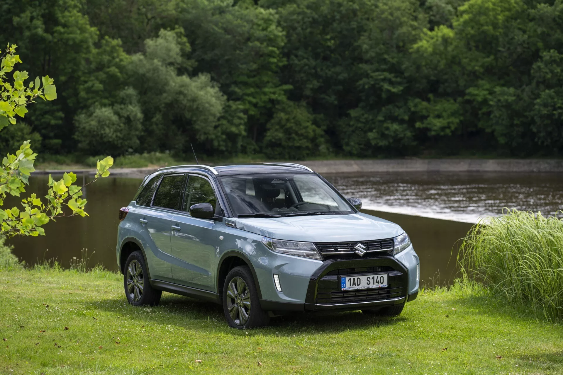 Photographie d'un SUV moderne Suzuki Vitara 2024 (2e génération, facelift 2) avec une carrosserie bicolore bleu clair et toit noir, garé sur un champ d'herbe avec de l'eau et des arbres en arrière-plan. Le SUV, vu d'un angle avant quart montrant le côté conducteur, présente une calandre avant proéminente avec un emblème central, des phares LED angulaires élégants, un capot lisse, des roues en alliage multibrins et une garde au sol notable. Les vitres teintées, les rétroviseurs latéraux et les poignées de porte de la couleur de la carrosserie mettent en valeur son design contemporain, tandis que les rétroviseurs latéraux intègrent des feux de signalisation LED.