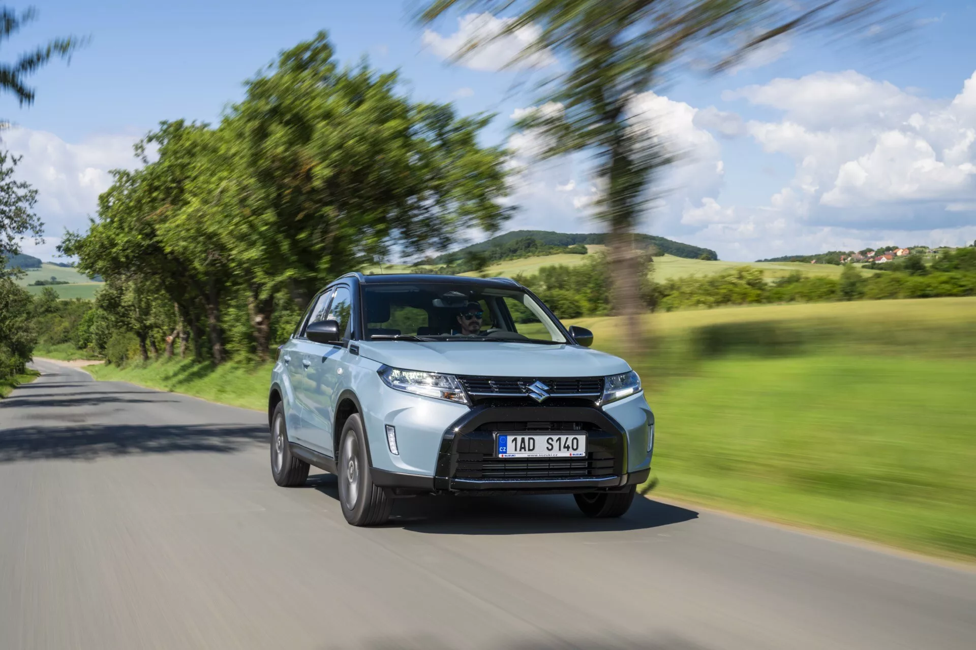 Le Suzuki Vitara 2024 2ème Génération Facelift est capturé en mouvement avec un fond flou de arbres et ciel, soulignant sa vitesse. Cette vue oblique avant met en valeur le SUV moderne avec une garde au sol élevée, une calandre distinctive, des lumières diurnes LED, des phares antibrouillard et des prises d'air. Les rétroviseurs latéraux, la carrosserie aérodynamique et les roues en alliage en mouvement soulignent son design. Les capteurs visibles suggèrent des systèmes avancés d'assistance à la conduite, comme le régulateur de vitesse adaptatif et l'évitement des collisions. La peinture métallique, l'apparence propre et la plaque d'immatriculation claire mais anonyme contribuent à son esthétique. Positionné sur une route de campagne, il montre la polyvalence du véhicule pour différents types de conduite.