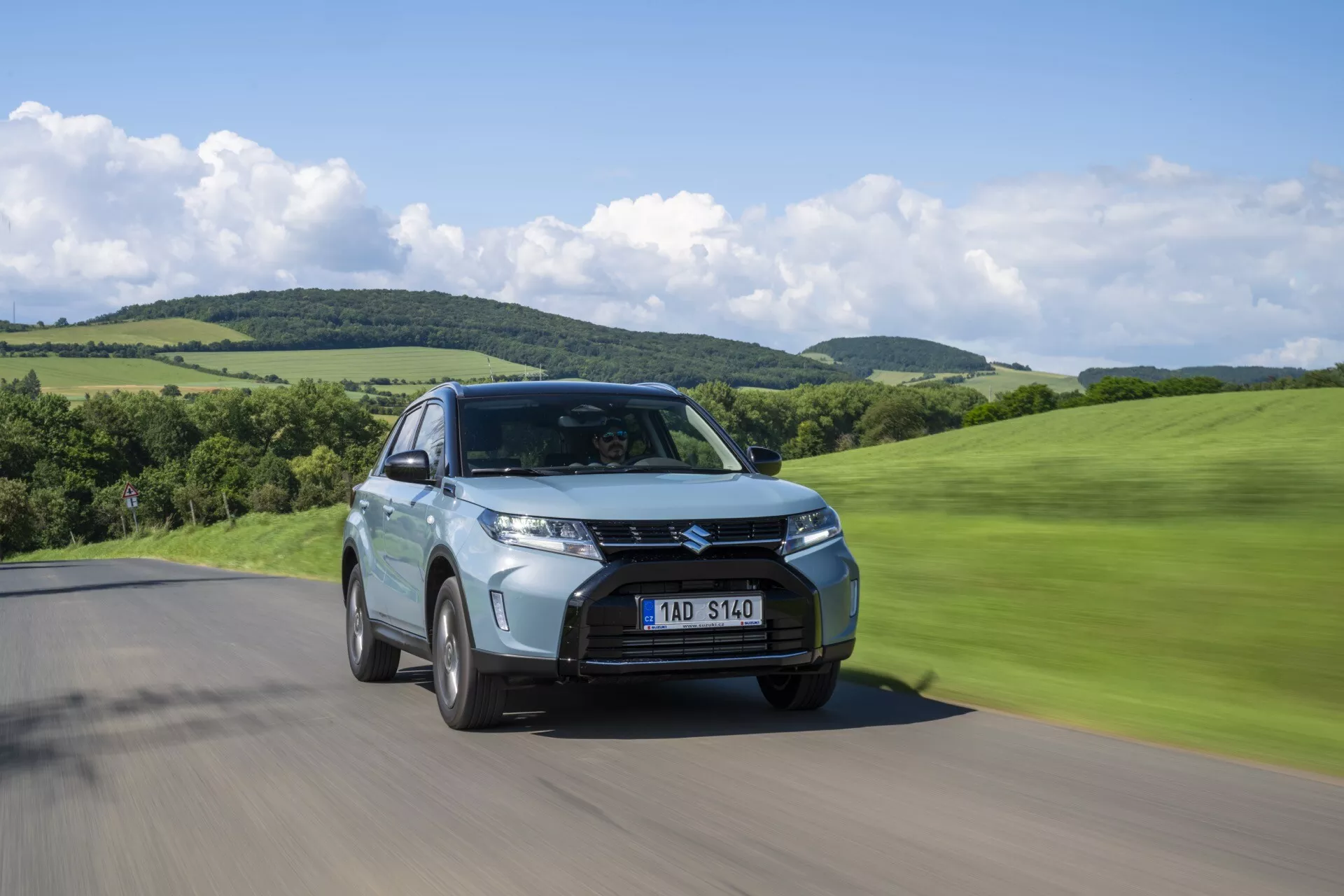 Vue en contre-plongée de la Suzuki Vitara 2024 restauration de deuxième génération sur une route, vue trois-quarts avant montrant la grille, les phares et le flou de mouvement dans un paysage vallonné vert.