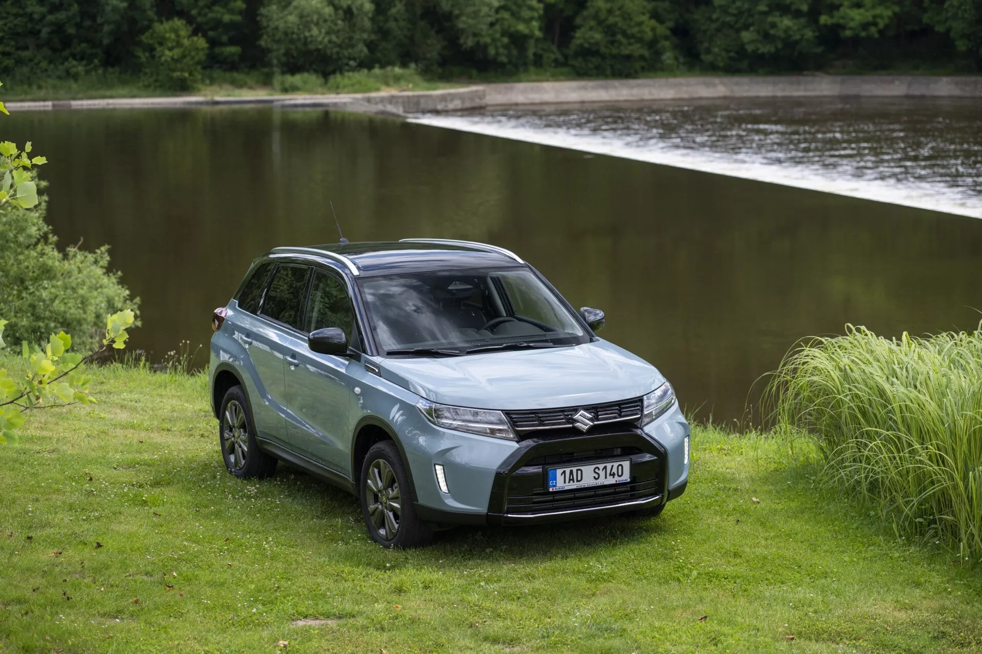 L'image présente le SUV Suzuki Vitara 2024 (seconde génération, second restylage) dans un cadre extérieur serein près d'un plan d'eau. Capturé en vue frontale trois-quarts, les phares LED élégants, la calandre et le pare-chocs avant sont bien visibles. Le capot sculpté et le pare-brise incliné ajoutent au profil aérodynamique. Un côté du SUV révèle un design à quatre portes, une zone de chargement arrière fermée, des jantes en alliage et des pneus tout-terrain robustes. La couleur argentée ou grise atténuée ajoute une touche de raffinement. Le terrain naturel et non pavé autour du SUV souligne ses capacités tout-terrain et son attrait aventurier.