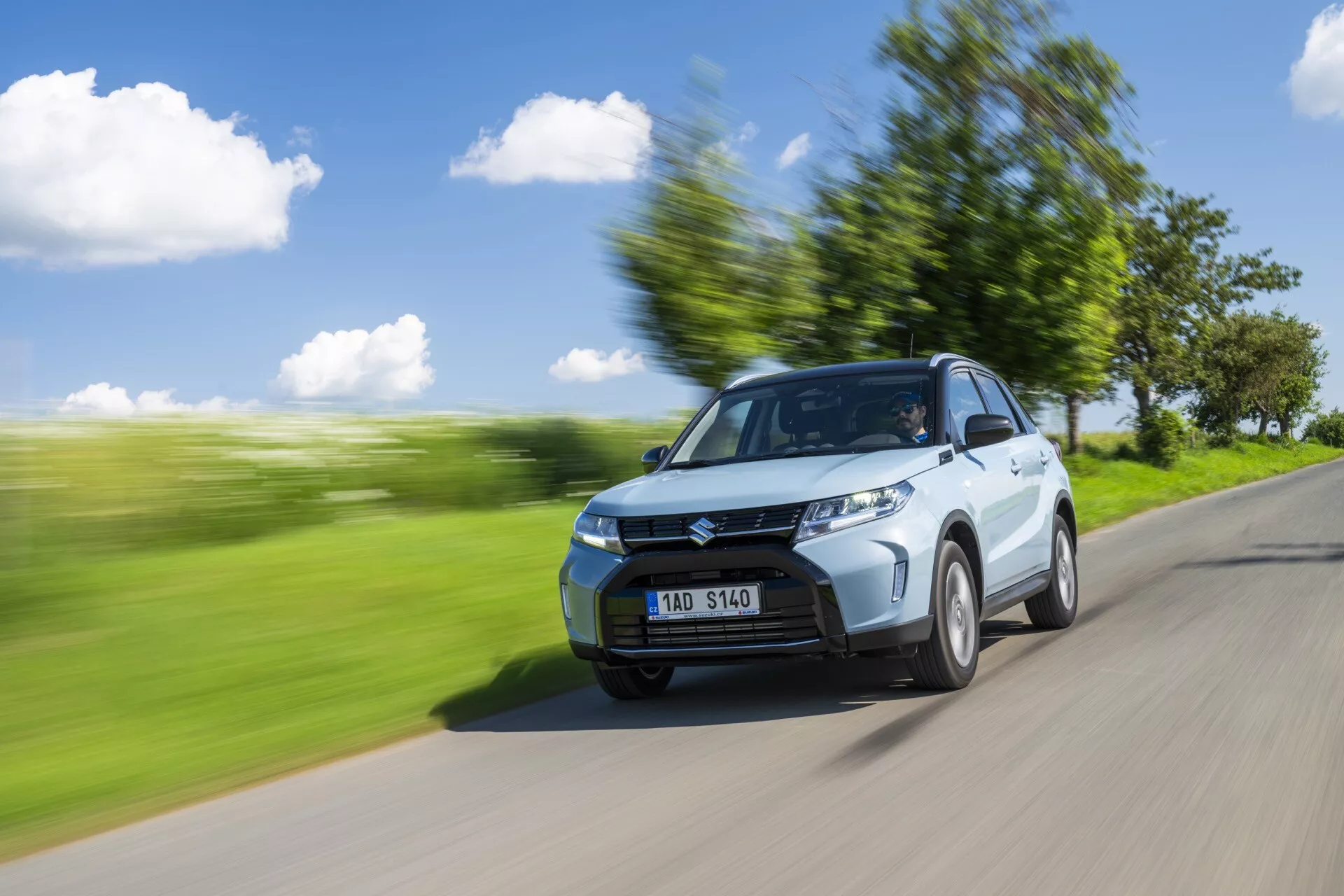 Prise de vue dynamique de la Suzuki Vitara 2024 (2e génération, lifting) capturée sous un angle trois-quarts avant. La voiture circule sur une route avec un paysage vert flou en arrière-plan, suggérant un mouvement. Les éléments visibles comprennent le capot, la calandre, les phares avant, le pare-chocs avant et une roue avant légèrement tournée. Le côté conducteur, y compris la porte et le rétroviseur latéral, est bien en vue. Le flou de mouvement et les traînées dans l'environnement accentuent la sensation de vitesse tandis que la voiture reste nette. Les lignes de contour, les découpes latérales et le design des roues sont évidents. Les conditions lumineuses diurnes avec un ciel clair et des ombres suggèrent une lumière solaire provenant du coin supérieur gauche. On aperçoit une personne à travers le pare-brise, probablement le conducteur.