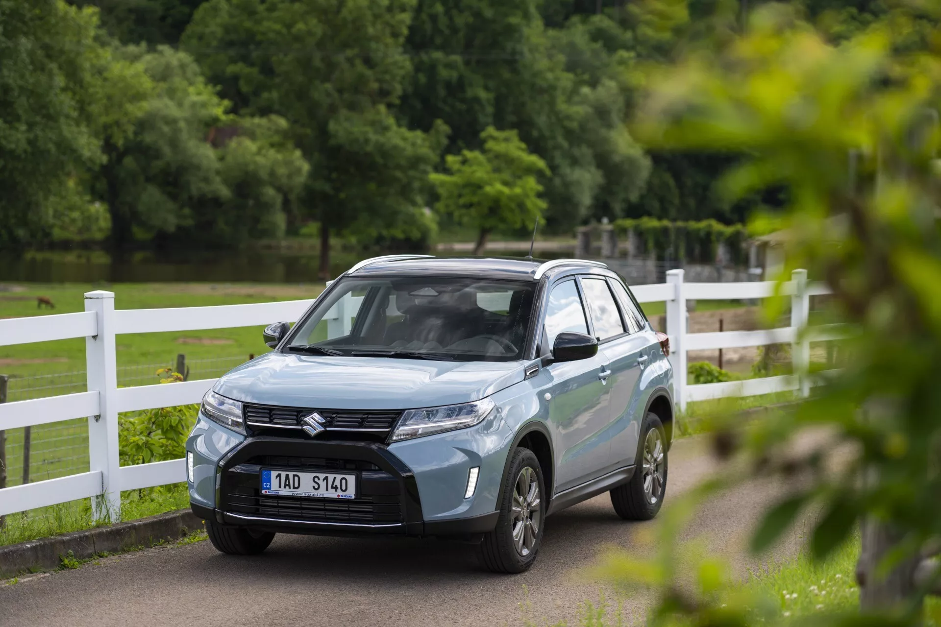 SUV Suzuki Vitara 2024 bleu clair dans un cadre de parc ou campagne, vu sous un angle avant trois-quarts montrant sa calandre sophistiquée avec emblème, phares LED acérés, lignes aérodynamiques du capot et roues en alliage à plusieurs branches. La plaque d'immatriculation avant du véhicule est floutée et les rétroviseurs latéraux pourraient intégrer des clignotants. La photo capture la voiture avec une faible profondeur de champ, mettant en valeur son design moderne tandis que l'arrière-plan reste légèrement flou.