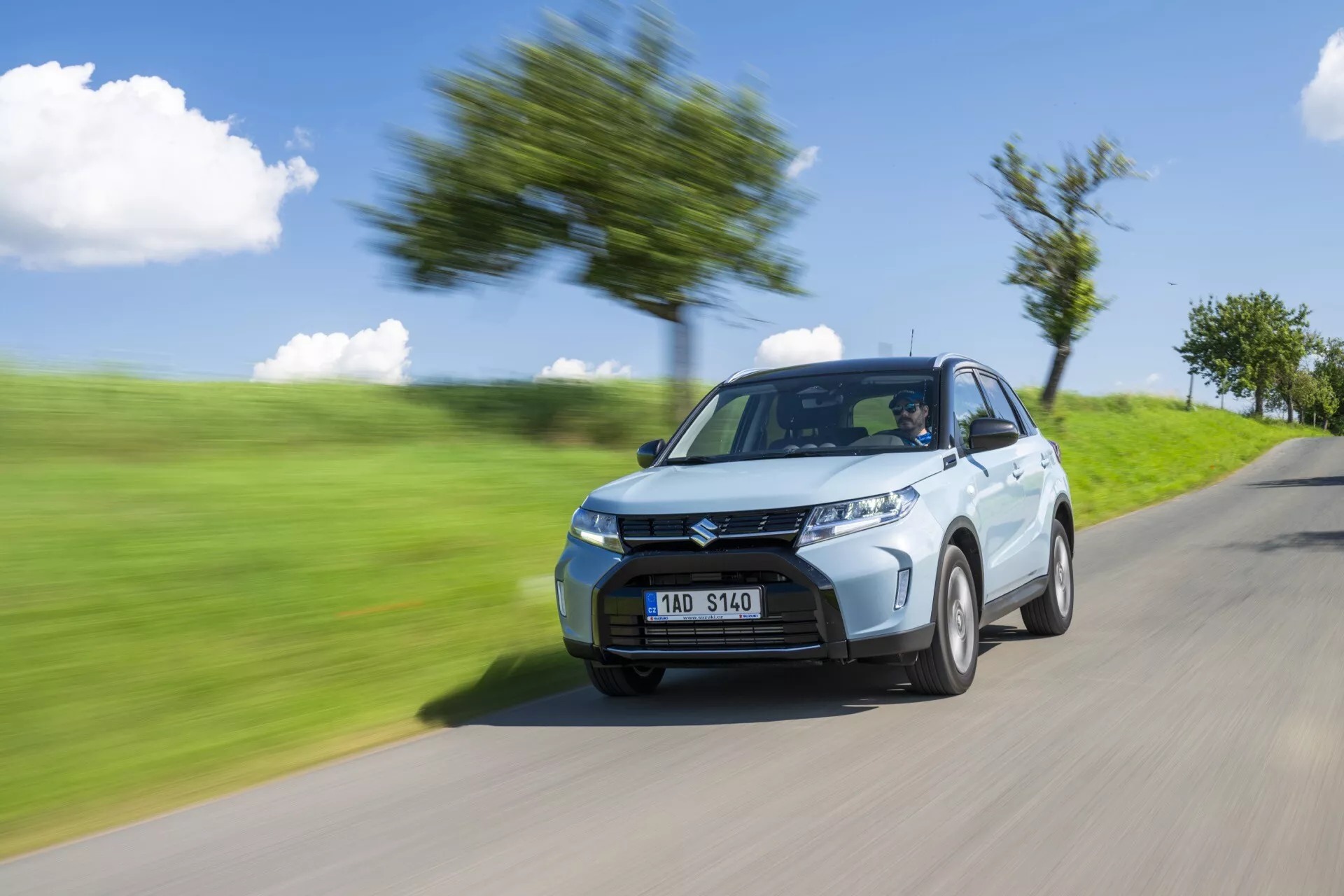 Prise dynamique du Suzuki Vitara 2024 (deuxième génération facelift), montrant son design moderne et sa vitesse. Capturé dans une perspective de quart avant-gauche, l'image met en valeur les phares tranchants, la calandre avant proéminente et les roues en alliage standard. L'effet de flou de mouvement sur l'arrière-plan et les roues accentue le mouvement rapide de la voiture, visible sur une route floue avec des arbres et de l'herbe.
