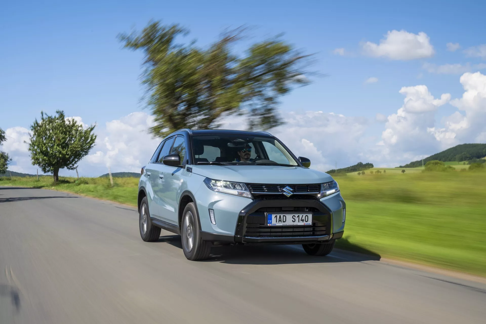Image dynamique de la Suzuki Vitara 2024 (2e génération, facelift 2) en mouvement, présentant l'angle trois quarts avant. La voiture est nettement focalisée, tandis que l'arrière-plan est flou en raison du mouvement, mettant en valeur ses phares, sa calandre proéminente, ses jantes en alliage, son style de carrosserie SUV compacte, sa peinture métallisée et son design aérodynamique. Idéal pour les présentations automobiles et les usages promotionnels.