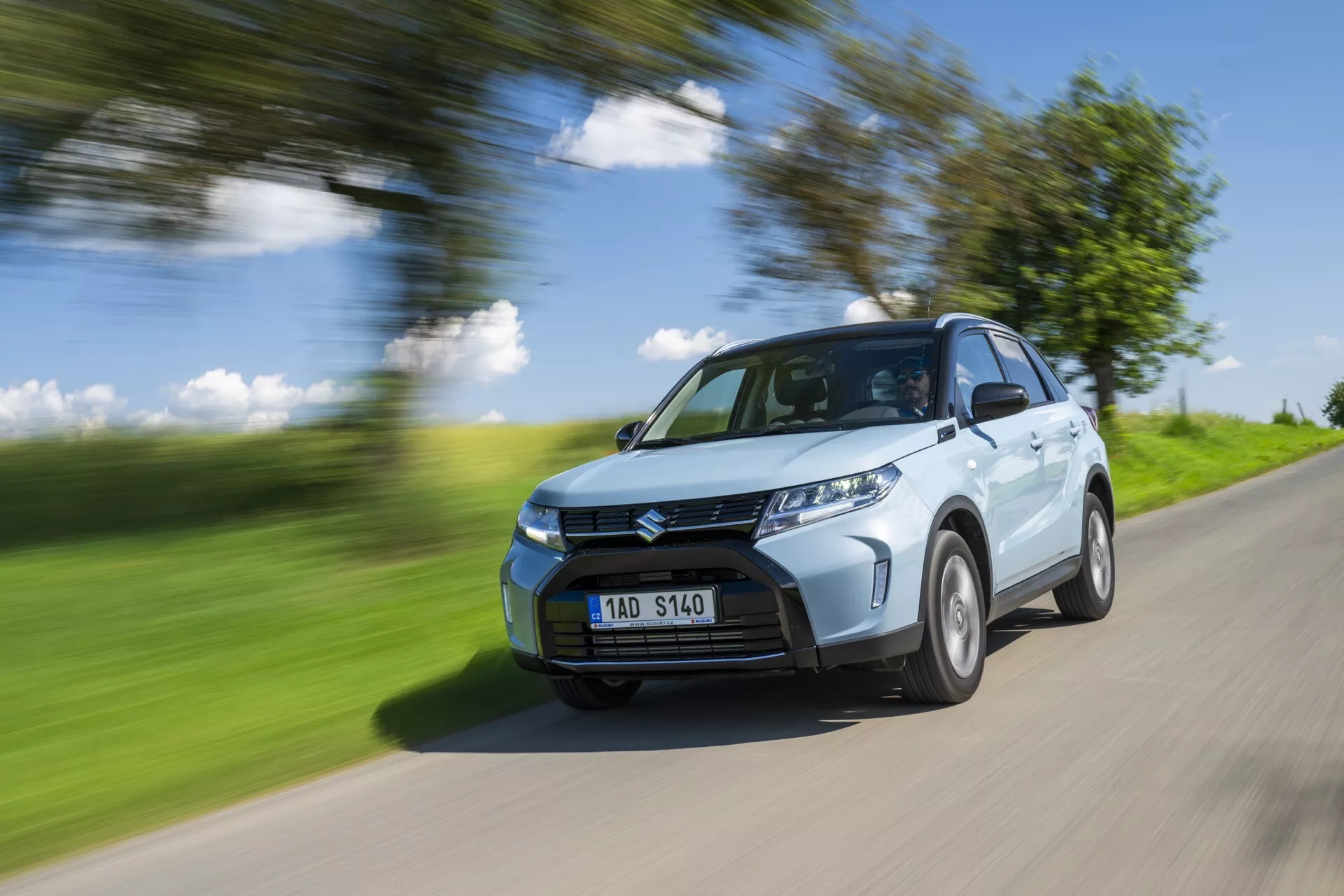 Un SUV compact Suzuki Vitara 2024 (restylage de 2e génération) bleu clair en mouvement sur une route de campagne. L'image capture le quart avant de la voiture, montrant la calandre avant, les phares, une partie du capot, le pare-brise et le côté conducteur, y compris l'arche de la roue avant, une partie de la porte arrière et une portion de l'arche de la roue arrière. La voiture est nette contre un arrière-plan et une route flous, indiquant la vitesse. Les roues montrent un léger flou suggérant la rotation. Éclairée par une lumière naturelle vive par une journée ensoleillée, projetant des ombres sur la partie inférieure du véhicule, l'image met en évidence les éléments de design tels que les plis du capot, le design du pare-chocs avant et le motif de la grille. Les phares LED sont visibles, indiquant potentiellement des fonctionnalités modernes comme des capteurs de stationnement. La plaque d'immatriculation est visible sans informations sensibles.