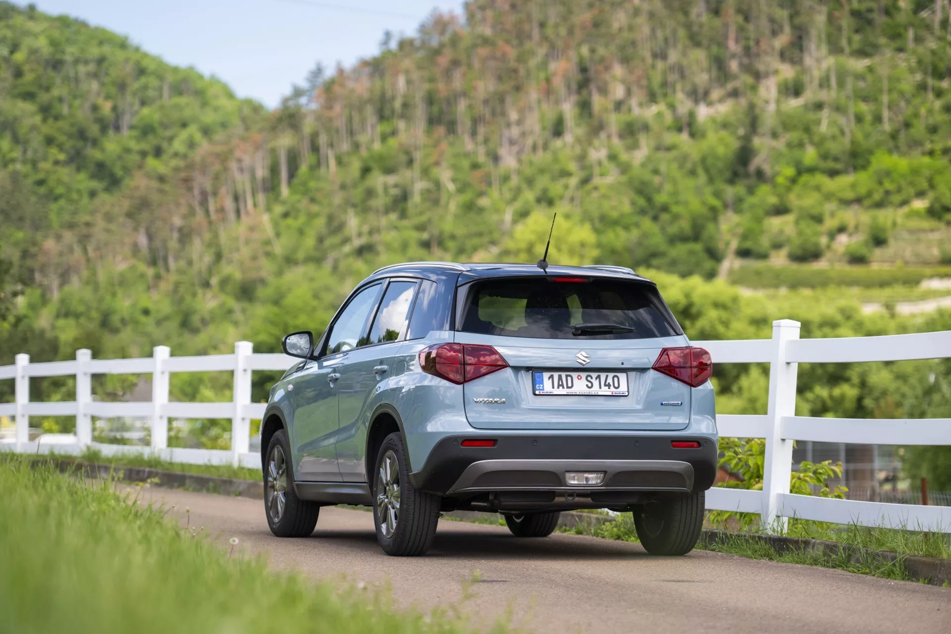 L'image montre la Suzuki Vitara 2024 (2e génération, restylée) en vue arrière trois-quarts, mettant en valeur le profil arrière droit tout en étant positionnée sur la route et légèrement inclinée à gauche. Les éléments visibles incluent le hayon, la lunette arrière, les portes et fenêtres partielles côté passager droit, les feux arrière à LED, les capteurs de stationnement sur le pare-chocs arrière, le spoiler de toit et une courte antenne de toit. La voiture est équipée de jantes en alliage et d'une plaque d'immatriculation visible. Elle se trouve dans un décor rural avec une clôture blanche et une végétation luxuriante.
