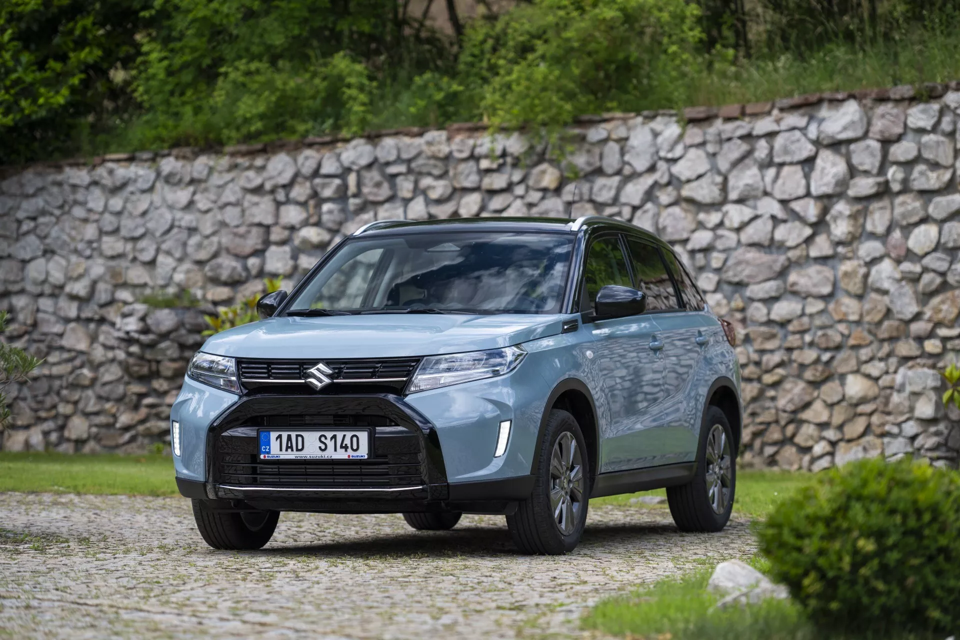 Photographie de haute qualité d'une voiture Suzuki dans un cadre extérieur, capturée sous un angle de trois quarts avant près de verdure ou d'un parc. La vue comprend la calandre avant avec un badge Suzuki proéminent, des phares au design moderne probablement dotés de la technologie LED, des antibrouillards intégrés dans le pare-chocs et le profil latéral côté conducteur avec les roues, la porte et les rétroviseurs visibles. La voiture repose sur une surface en gravier avec un mur de pierre en arrière-plan, accentuant l'ambiance naturelle et rustique. La scène à éclairage doux, probablement couverte, présente une faible profondeur de champ, floutant l'arrière-plan pour concentrer l'attention sur les détails du design et technologiques de la voiture.