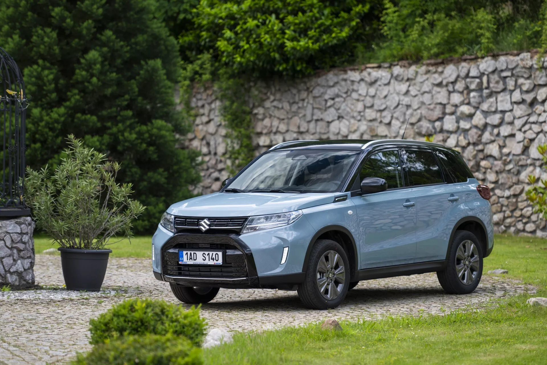 Vue latérale avant en angle d'un SUV moderne Suzuki Vitara 2024 stationné à l'extérieur dans un quartier résidentiel ou un parc avec une pelouse bien entretenue et un mur de pierre en arrière-plan. L'image, prise à une distance moyenne, montre la calandre avant avec des barres horizontales arborant le logo de la société, des phares à LED élégants avec feux de jour, un pare-chocs sculpté avec phares antibrouillard intégrés, des roues en alliage, des rétroviseurs latéraux avec clignotants intégrés, une finition de peinture métallique, des lignes dynamiques de la carrosserie, des vitres teintées, des rails sur le toit et une antenne sur le toit, mettant en valeur son design et ses caractéristiques modernes.