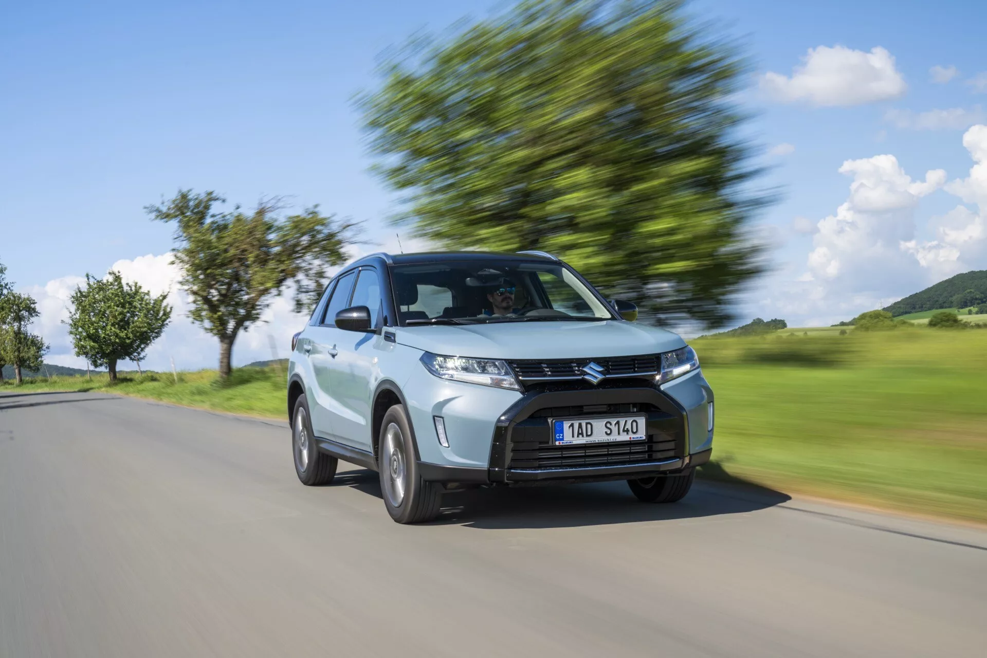 Image dynamique d'une voiture hatchback argentée Suzuki Vitara 2024 en mouvement, capturée sous un angle frontal trois-quarts, mettant en évidence son design moderne avec une calandre proéminente, des phares angulaires, un toit incliné et des roues en alliage en mouvement avec un flou d’arrière-plan signifiant vitesse.