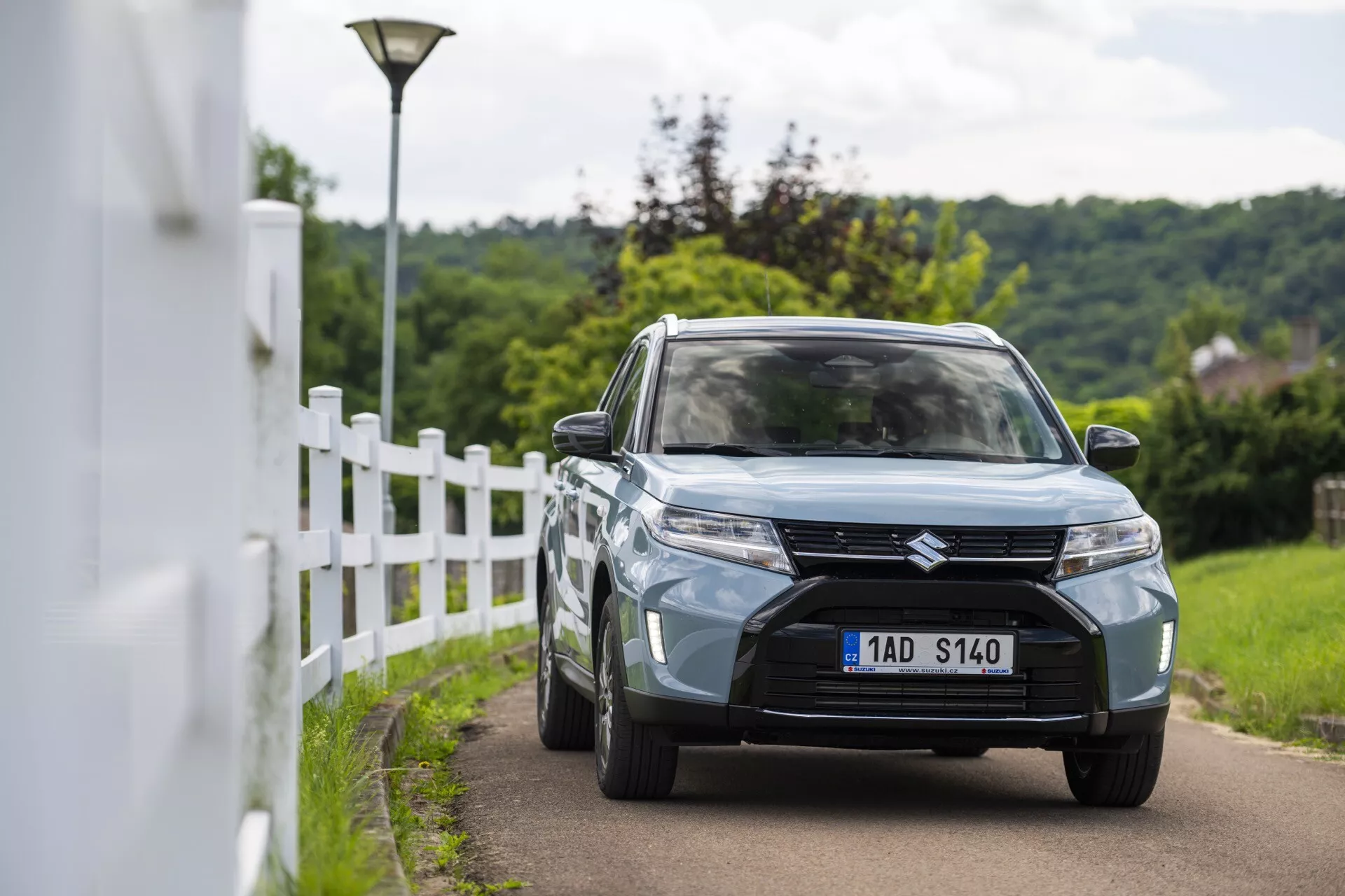 Scène en plein jour à l'extérieur représentant la Suzuki Vitara 2024 (restylage de la 2ème génération) capturée sous un angle de vue trois quarts avant. L'image met en avant les éléments de design du véhicule, y compris la calandre avant proéminente avec un design à lamelles horizontales et un badge central, des phares modernes à LED ou HID, des feux antibrouillard encastrés dans la partie inférieure du pare-chocs, et une couleur bleu clair ou gris. La voiture roule sur une voie étroite près d'une clôture blanche avec un arrière-plan légèrement flou, suggérant une faible profondeur de champ. Les autres caractéristiques visibles comprennent des roues en alliage, des rétroviseurs latéraux avec clignotants intégrés et un pare-chocs avant partiellement visible avec une prise d'air centrale.