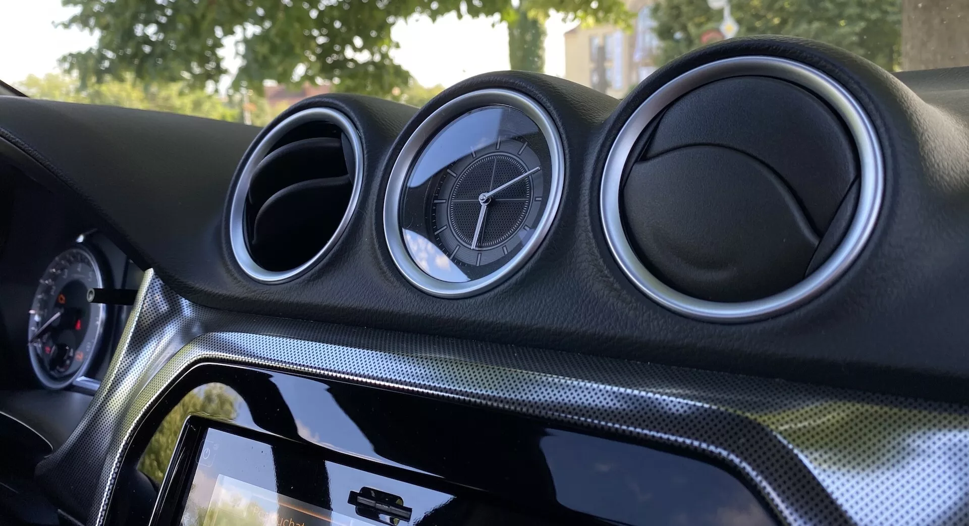 Vue intérieure en gros plan de la Suzuki Vitara 2018 se concentrant sur le tableau de bord central. Le tableau de bord présente trois aérateurs circulaires métalliques avec l'emblème du fabricant, un système d'infodivertissement à écran tactile avec un cadre noir brillant, un matériau à apparence en fibre de carbone et un éclairage ambiant pour améliorer l'esthétique.