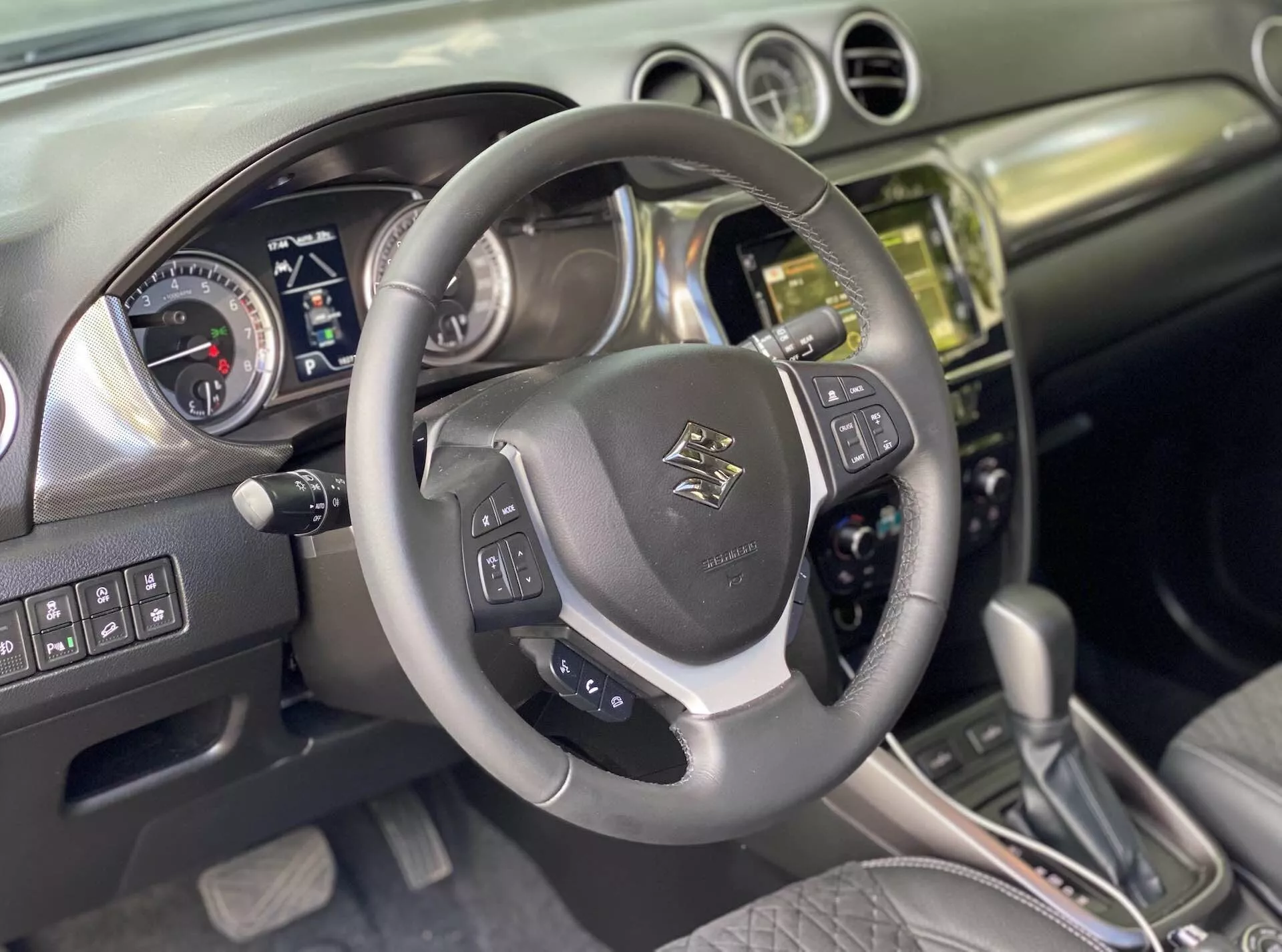 Intérieur du Suzuki Vitara 2018, capturant la perspective du conducteur. Gros plan sur un volant multifonction en cuir avec emblème au centre, commandes montées pour l'audio, le contrôle vocal et le régulateur de vitesse avec accents métalliques. Derrière, un tableau de bord avec cadrans analogiques pour le compteur de vitesse et le compteur de régime, et un écran digital pour les informations sur le véhicule, suggère que la voiture est éteinte ou en mode accessoire. La console centrale présente une pile finie en métal ou chrome avec un système infotainment à écran tactile, des boutons et des commandes pour le climat et le véhicule. Le levier de transmission automatique avec accents en cuir suggère plusieurs modes de conduite. L'intérieur combine cuir et métal brossé, reflétant un design moderne. On déduit des fonctionnalités avancées comme le Bluetooth, la navigation, les assistances au stationnement et les systèmes d'assistance au conducteur.