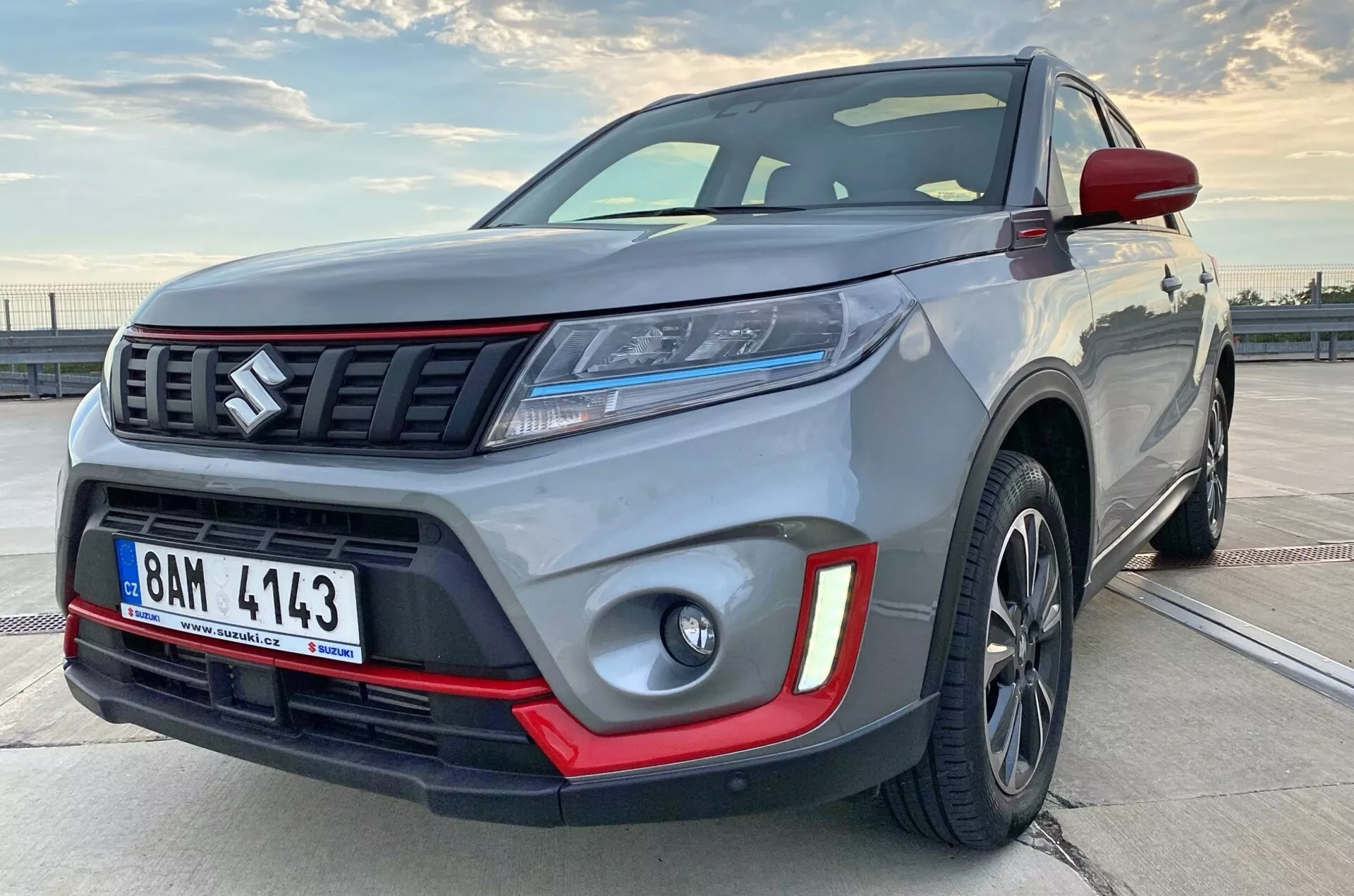 Vue latérale frontale rapprochée d'un SUV compact moderne, inclinée pour montrer une partie du profil latéral. L'image met en évidence la robustesse du véhicule avec des détails visibles tels que le pare-chocs avant, la calandre avec des barres horizontales et un emblème, des phares LED élégants, des phares antibrouillard avec inserts rouges, des roues en alliage et des ailes musclées. Les roues en alliage foncé et la pièce de garniture contrastante suggèrent une version premium ou sportive.