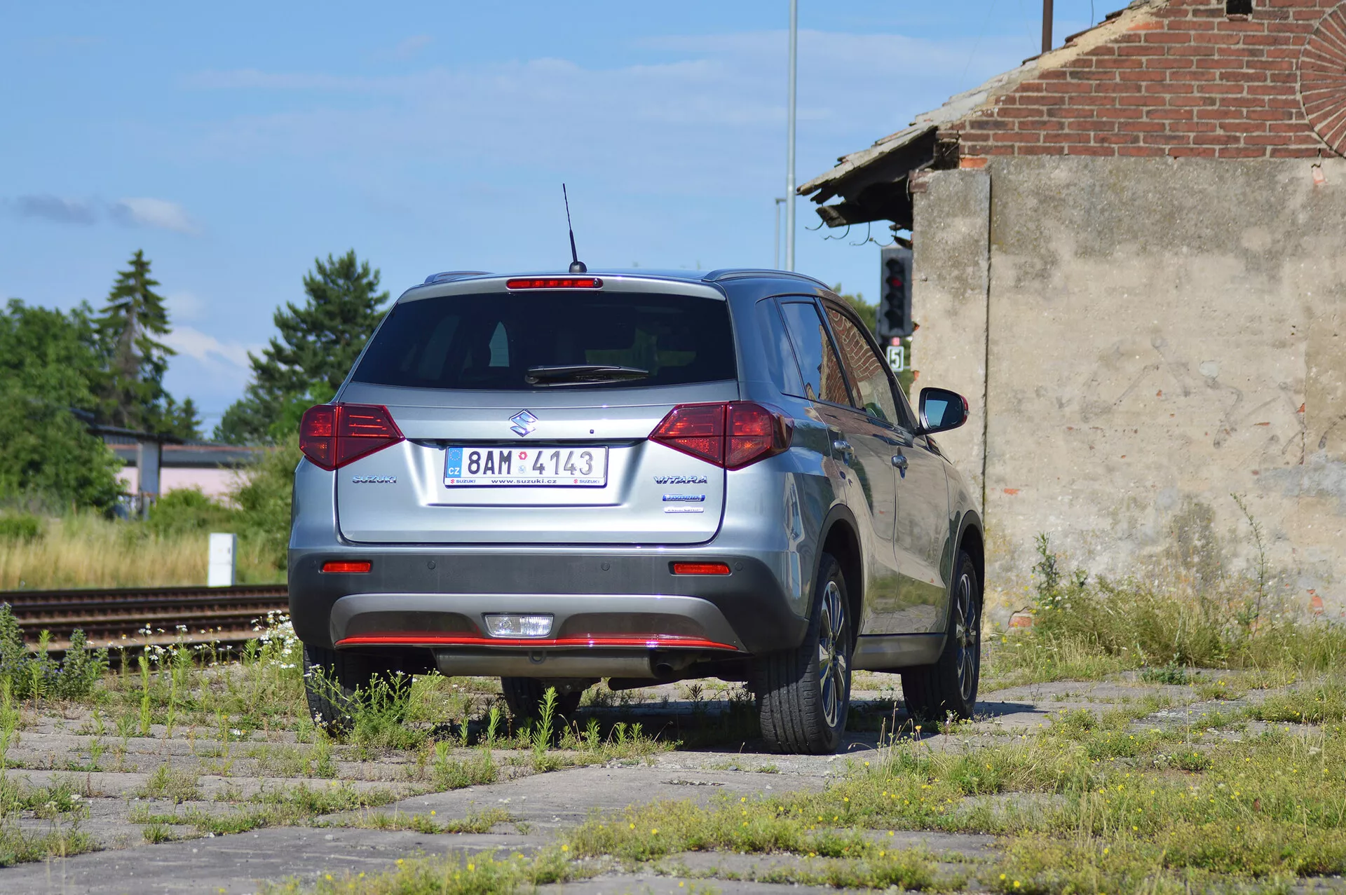 Un SUV compact Škoda Yeti gris métallisé stationné sur un terrain négligé avec des plaques d'herbe et de béton, photographié d'une vue arrière trois-quarts. L'image montre le design arrière distinctif du véhicule avec des feux arrière conventionnels, un essuie-glace arrière, un feu stop monté en hauteur et une partie d'un becquet arrière. Le logo Škoda est centré sur le hayon, le nom du modèle 'Yeti' à gauche et un badge '4x4' à droite, indiquant une transmission intégrale. La voiture a des jantes en alliage à cinq branches, semble en bon état et est située dans une zone industrielle avec des voies de chemin de fer désaffectées et une structure délabrée en arrière-plan, sous une lumière naturelle.