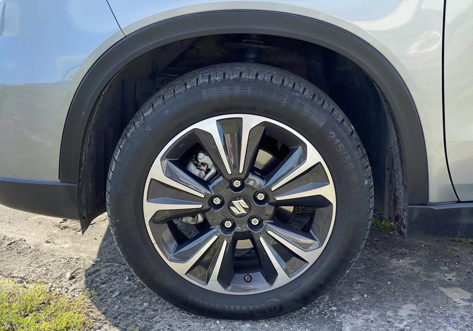Vue rapprochée de la roue de la Suzuki Vitara 2018, présentant un design en alliage avec plusieurs rayons métalliques et des zones peintes plus foncées. Le capuchon central marqué cache les écrous de roue, et le pneu pour route affiche des motifs de bande de roulement détaillés et des marquages sur le flanc. L'arrière-plan comprend une portion de l'aile et du panneau latéral du véhicule de couleur neutre et brillante, vue d'un léger angle vers le haut depuis le sol.