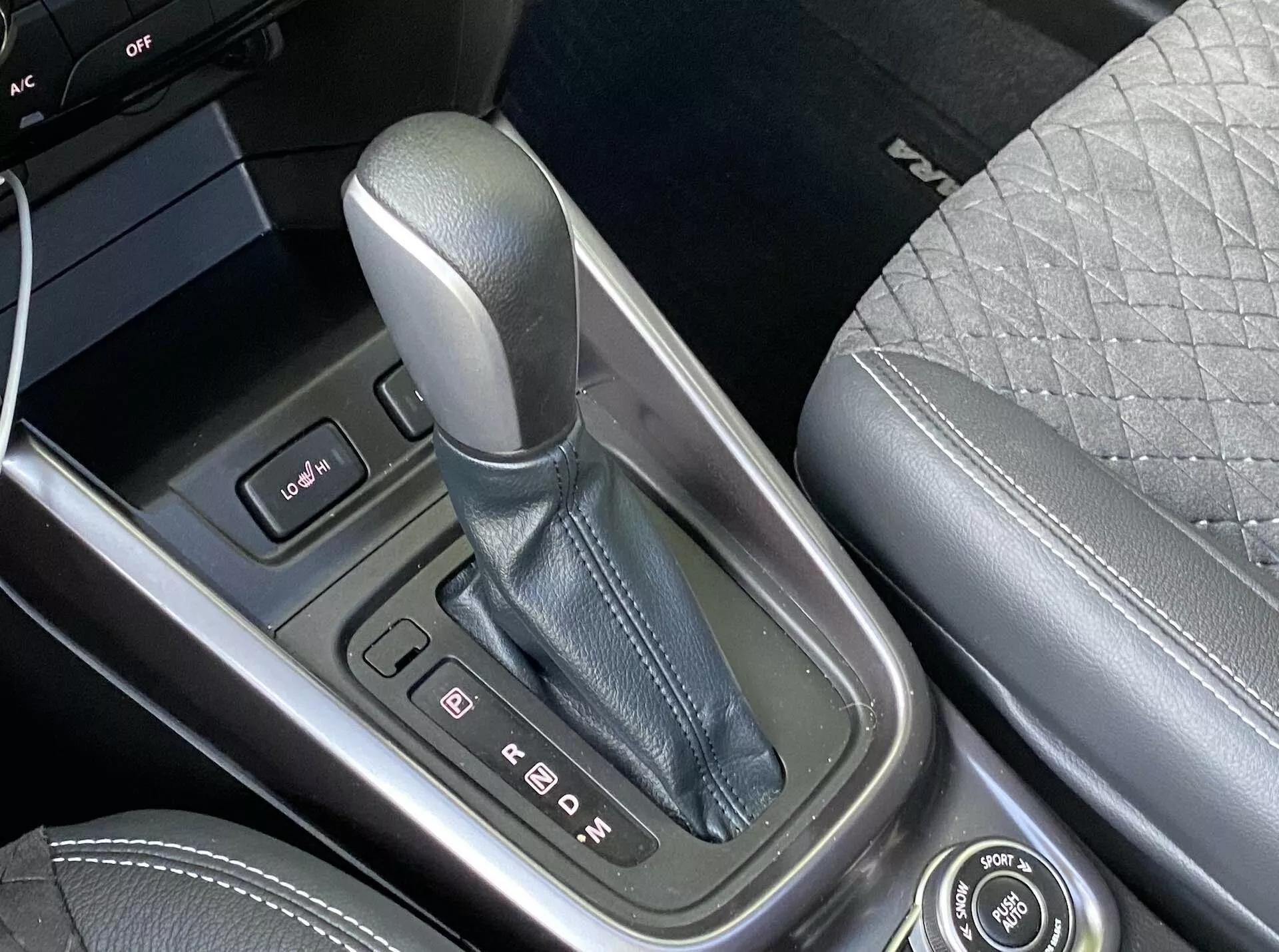 Vue rapprochée de la console centrale de la Suzuki Vitara 2018, mettant l'accent sur le levier de vitesse en position 'P' pour le stationnement, entouré d'un boîtier à finition métallique avec des indicateurs de vitesse clairs. À gauche, on voit un panneau de commande avec deux boutons et le bord d'un écran. La console centrale est fabriquée avec des matériaux de haute qualité et à droite, on peut distinguer une partie du siège passager avant avec un design matelassé. Un bouton rotatif pour les fonctions d'infodivertissement est également présent, soulignant la technologie avancée et les caractéristiques de luxe du véhicule.