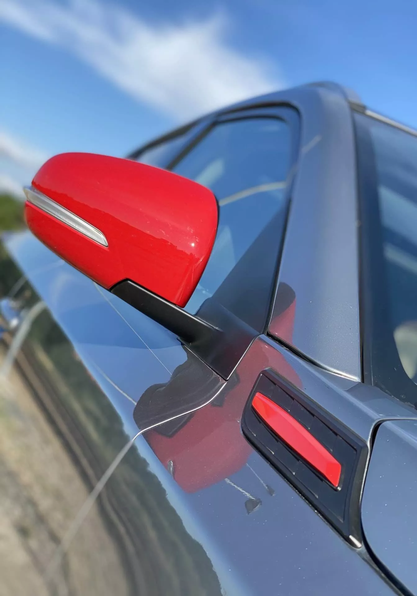 Vue en gros plan du rétroviseur côté conducteur d'une Suzuki Vitara 2018, mis en évidence en rouge contre la carrosserie sombre de la voiture. Le rétroviseur est fixé avec un support noir élancé, suggérant une efficacité aérodynamique. La prise de vue capture le rétroviseur sous un angle oblique légèrement avancé, avec une perspective orientée vers le bas, présentant un bandeau rouge assorti sur la porte. L'arrière-plan est flou, mettant en valeur les détails du rétroviseur et du design de la porte de la voiture.
