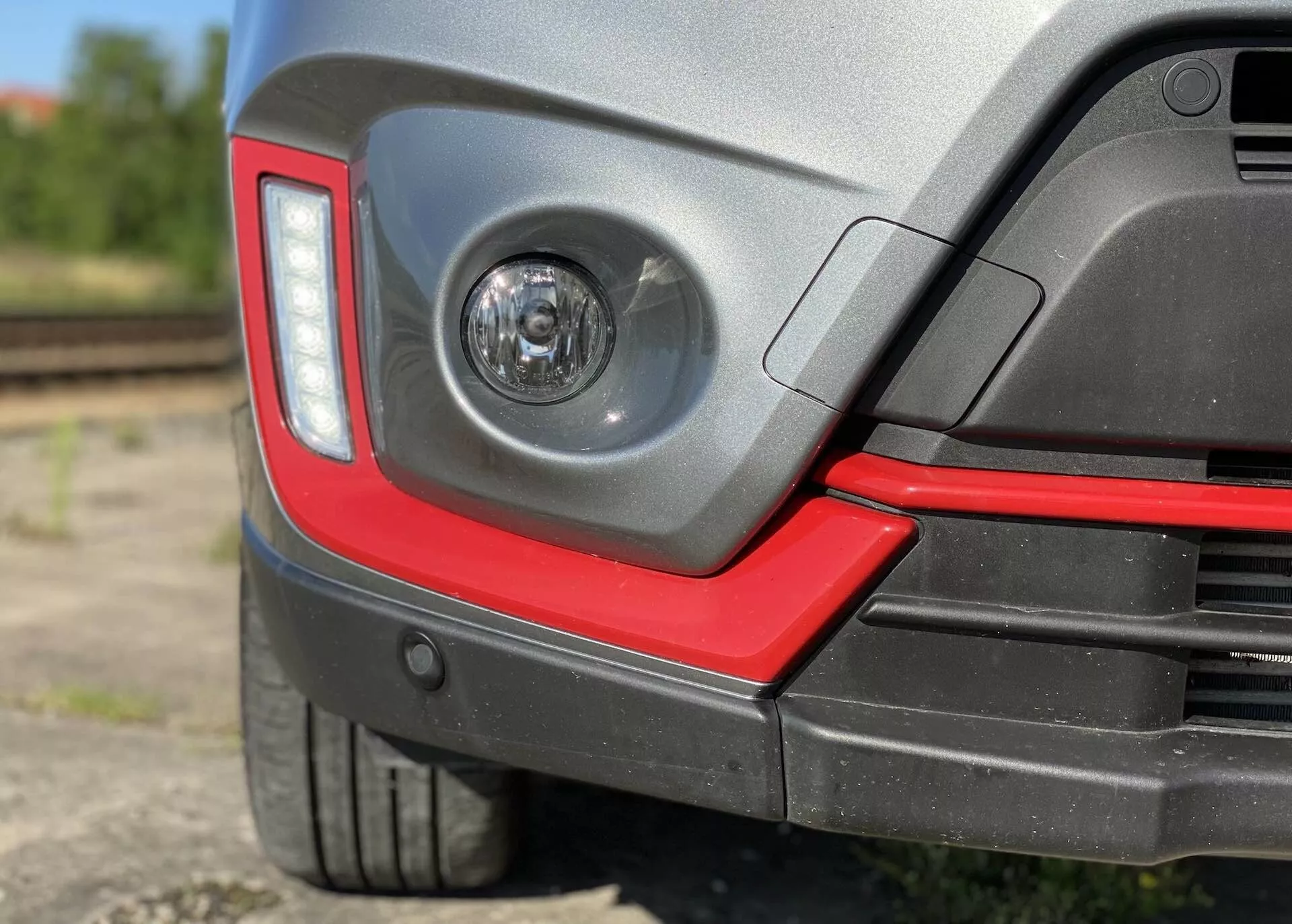 Vue rapprochée de la section d'angle arrière d'une Suzuki Vitara 2018, présentant un feu antibrouillard à lentille claire dans le pare-chocs arrière, un design moderne et angulaire des feux arrière, un pare-chocs arrière bicolore avec un accent rouge sportif, des capteurs de stationnement potentiels, un pneu à profil bas avec un passage de roue en plastique protecteur, une finition de peinture brillante et un arrière-plan flou semi-industriel avec des voies ferrées.