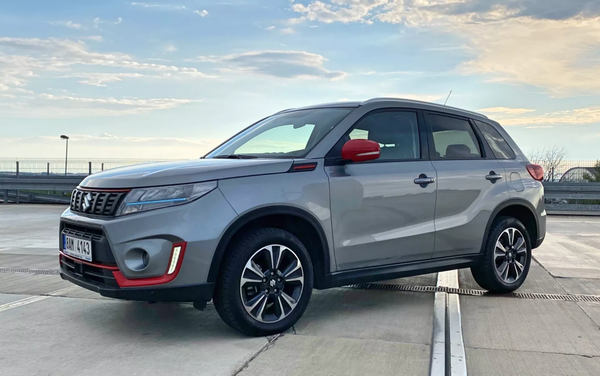 L'image montre un Suzuki Vitara 2018, un SUV compact moderne, garé en extérieur en pleine journée. Il est capturé sous un angle trois-quarts avant, mettant en avant les profils avant et latéral. L'angle est légèrement bas, au niveau des poignées de porte, ce qui suggère que le photographe était accroupi ou penché. Les détails visibles incluent la calandre avant, les phares, le boîtier des feux anti-brouillard, l'emblème, les rétroviseurs latéraux, les jantes en alliage avec clignotants intégrés, les poignées de porte, les garnitures de fenêtre, la ligne de toit et les rails de toit. Les phares à projection et l'antenne élégante suggèrent des technologies avancées. Les éléments de design moderne du SUV indiquent des fonctionnalités telles que les feux LED et possiblement un système d'entrée sans clé.