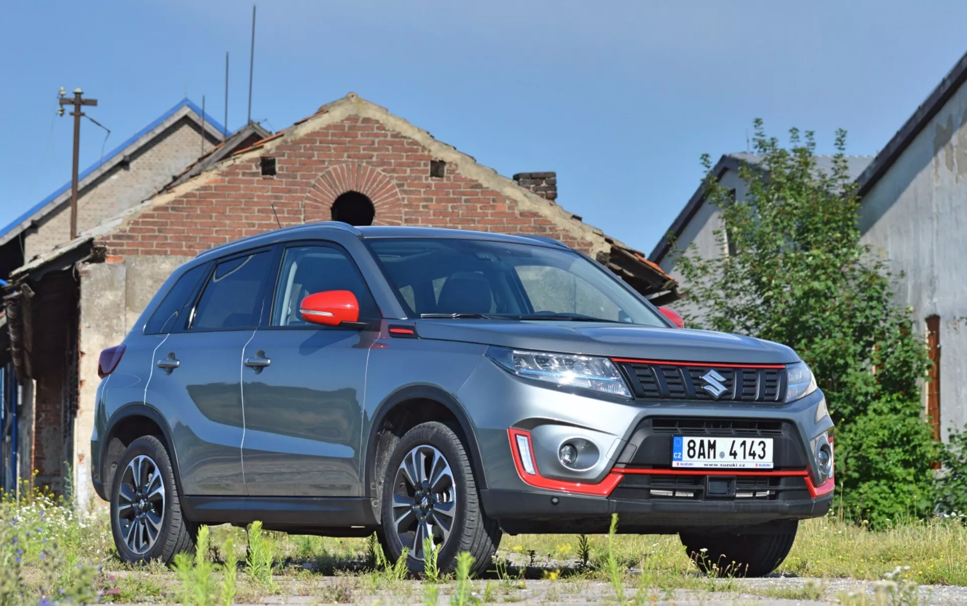 L'image montre un SUV Suzuki Vitara 2018 sous un angle de vue avant trois quarts bas, mettant en évidence le côté avant droit du véhicule avec une garde au sol notable. Les caractéristiques visibles incluent un pare-chocs avant robuste avec des feux de brouillard et une plaque de protection argentée, un emblème de marque sur la grille à lamelles horizontales, des lignes de capot définies, des phares modernes possiblement avec technologie LED ou HID, des jantes en alliage multirayons, des rétroviseurs latéraux rouges avec clignotants intégrés, des ailes musclées, un pare-brise réfléchissant et des vitres teintées. Le Vitara est garé sur une surface en herbe non pavée, devant un vieux bâtiment ajoutant un arrière-plan industriel ou rural, suggérant une aptitude pour les environnements extérieurs.