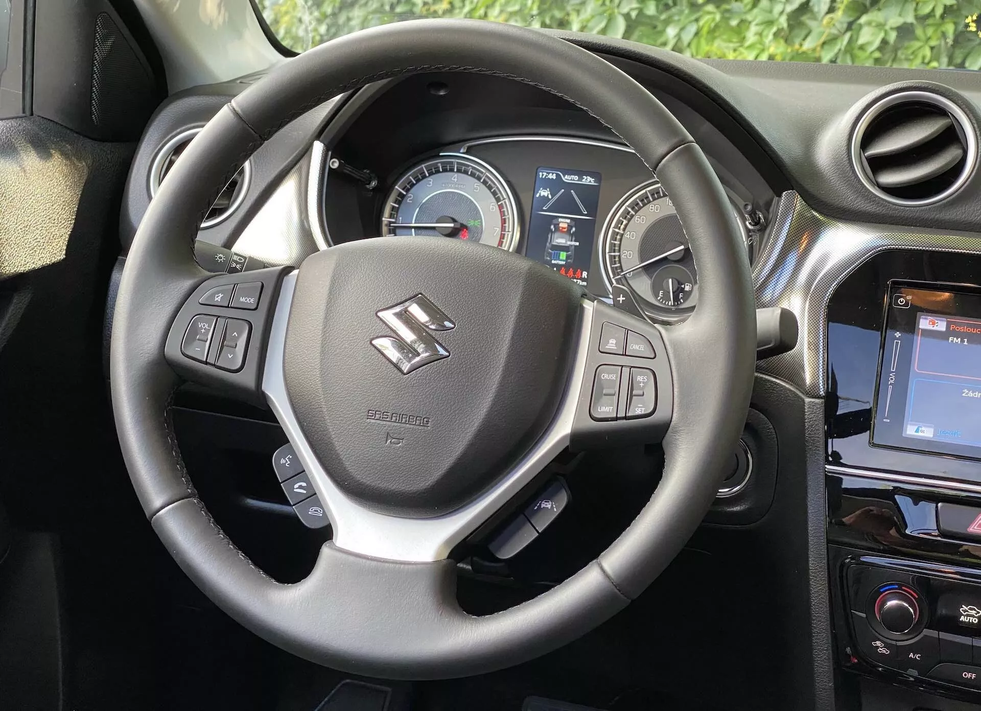 Image de l'intérieur d'une Suzuki Vitara 2018, mettant en avant la perspective du conducteur avec un focus sur le volant et le tableau de bord. Le volant, centré dans l'image, est doté de repose-pouces et de commandes pour l'audio, les commandes vocales et le régulateur de vitesse. Derrière, un tableau de bord affiche un speedomètre analogique et partiellement un écran numérique fournissant des informations au conducteur. Une portion de la console centrale à droite révèle un système d'infodivertissement tactile, renforçant l'esthétique moderne de la voiture.