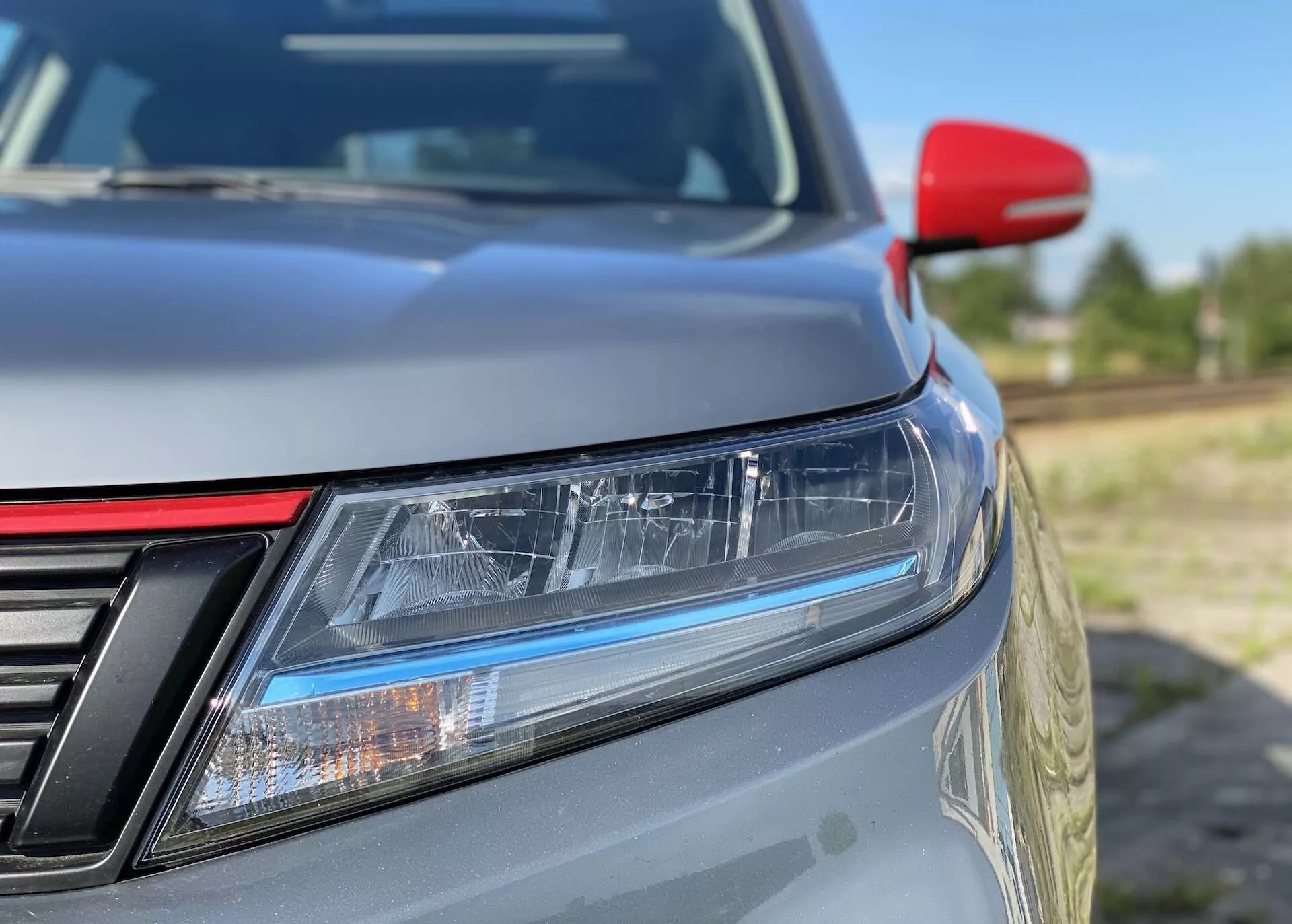 Gros plan de l'angle avant gauche d'une Suzuki Vitara 2018, mettant en avant des caractéristiques détaillées telles que des phares LED avec feux de jour, une grille partiellement visible avec des barres verticales, un capot légèrement incliné, une peinture métallisée, un rétroviseur latéral rouge contrastant probablement avec des clignotants intégrés et un arrière-plan flou qui accentue le design élégant et l'efficacité aérodynamique.