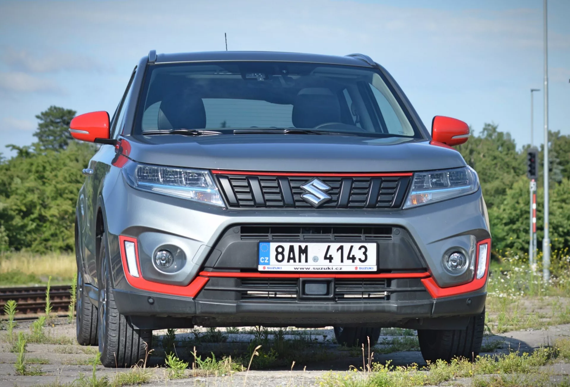 L'image montre un SUV Suzuki Vitara 2018 moderne sous un angle avant légèrement incliné, mettant en évidence la façade avant et une partie du côté passager sur un fondu de paysage rare avec des voies ferrées. La voiture, stationnée sur ce qui semble être une surface de gravier, présente une calandre proéminente avec un design angulaire, des ensembles de phares élégants probablement équipés de la technologie LED ou HID, et des feux de jour intégrés ou des phares antibrouillard dans le pare-chocs avant. Le pare-chocs comprend un insert de couleur contrastante, suggérant un niveau de finition tout-terrain ou sportif, et la hauteur de caisse visible renforce son caractère de SUV. Les lignes surélevées sur le capot contribuent à un style agressif, tandis que les rétroviseurs latéraux et les passages de roue montrent un design contemporain. Les jantes en alliage partiellement visibles complètent l'aspect robuste. La photo, prise d'une courte distance, permet un examen détaillé de ces caractéristiques.