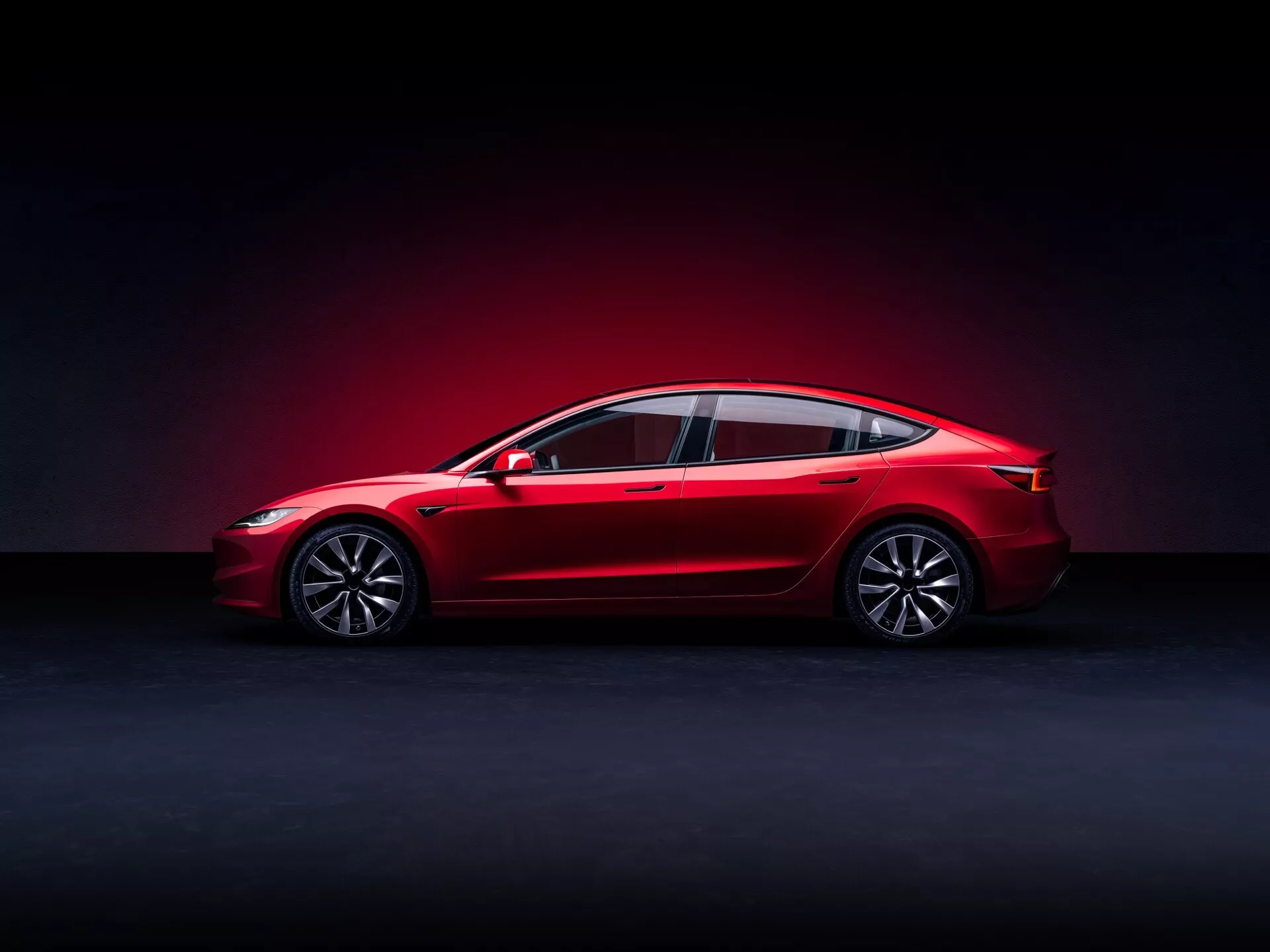 Tesla Model 3 2023 (1ère génération - Facelift) présentée de profil, soulignant son design élégant, visibilité complète incluant les roues avant et arrière, les vitres latérales et les rétroviseurs, avec un accent sur les lignes aérodynamiques, les roues en alliage et les caractéristiques technologiques modernes, le tout mis en valeur par un éclairage dramatique pour accentuer la finition rouge brillante