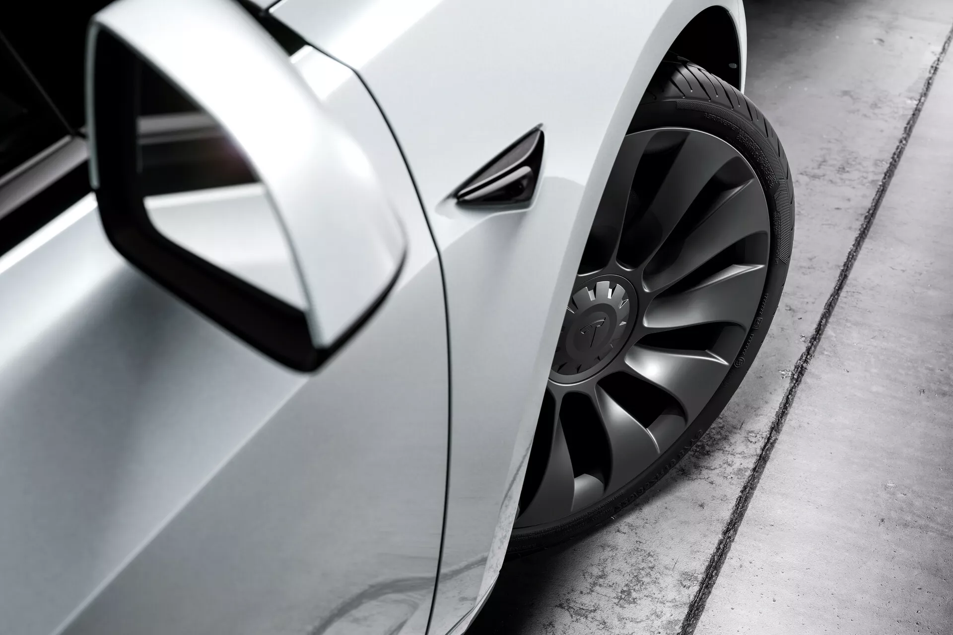 Vue latérale en gros plan d'une Tesla Model 3 argentée (2019, 1ère génération) centrée sur la roue avant, la zone de la portière conducteur et le rétroviseur latéral. La photo, prise depuis un angle bas proche du niveau du sol, met en valeur la grande jante en alliage au design multibrins foncé et le pneu à profil bas, le panneau latéral aérodynamique avec une finition métallisée et le rétroviseur latéral aérodynamique. La poignée de porte semble affleurante avec la carrosserie, suggérant une aérodynamique avancée, tandis qu'une petite ventilation derrière la roue avant pourrait servir au refroidissement des freins, à l'aérodynamique ou à l'esthétique. Le reflet bien défini sur le panneau indique une peinture de haute qualité.