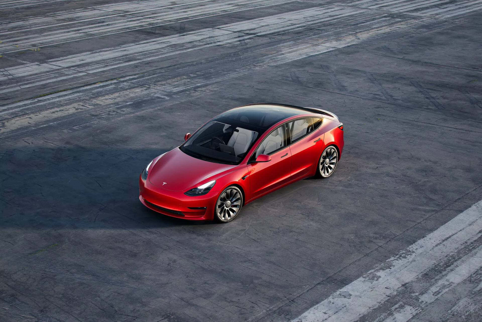 Prise de vue en plongée d'une berline Tesla Model 3 (2019, 1ère génération) rouge à quatre portes, garée sur une surface en asphalte texturé avec des marques de pneus visibles, suggérant une activité antérieure. La voiture est positionnée légèrement à droite du centre du cadre et est vue sous un angle de trois quarts avant, montrant le côté du conducteur avant et une vue partielle arrière. Le design aérodynamique, avec une ligne de contour distinctive, un toit panoramique en verre, des roues en alliage à plusieurs rayons, des phares à LED et une grille avant minimaliste, souligne la technologie avancée et le design électrique du véhicule.
