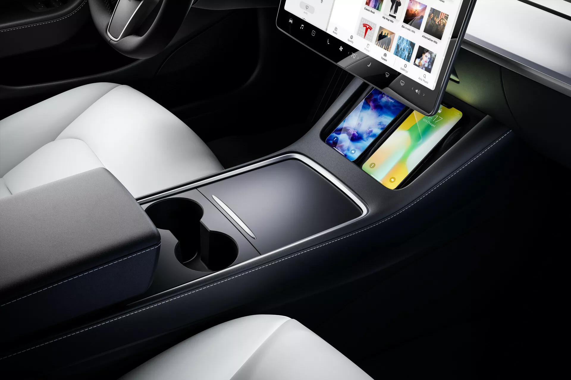 Vue rapprochée de l'intérieur du Tesla Model 3 2019, mettant en avant la console centrale et les sièges avant. L'image, prise d'un angle supérieur et diagonal, met en évidence le design épuré et minimaliste de la console centrale avec un grand écran tactile orienté à l'horizontale. Le système d'infodivertissement présente des options multimédia telles que l'intégration de smartphone et la navigation GPS. Sous l'écran tactile se trouve une surface plate, deux porte-gobelets et un compartiment de rangement ouvert. Les tons sombres de la console contrastent avec les sièges en cuir clair, reflétant un design luxueux et contemporain. En bas à gauche, on entrevoit une roue de contrôle, ajoutant à l'ambiance technologique de l'intérieur moderne de la voiture.