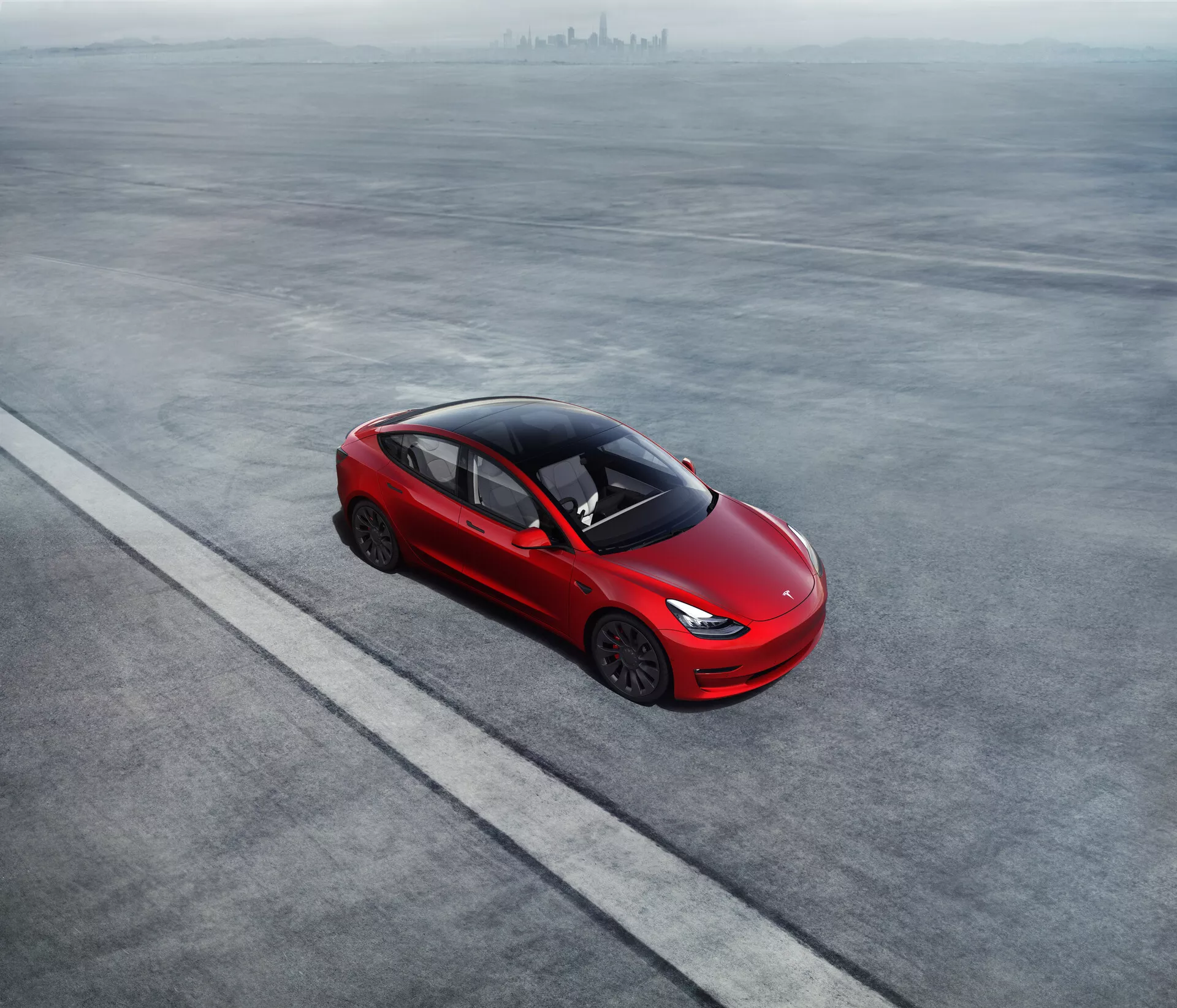Vue aérienne d’une Tesla Model 3 2019 (1ère Génération) rouge vif, garée sur une surface grise et plane. La berline quatre portes présente une silhouette aérodynamique, une ligne de toit inclinée se fondant dans le coffre et des roues en alliage uniques. Les poignées de portes encastrées suggèrent une efficacité aérodynamique. Le fort degré de teinte des vitres, l'absence de pot d'échappement visible et des phares fins et modernes impliquent une technologie avancée et potentiellement une propulsion électrique ou hybride. La voiture est positionnée parallèlement à une ligne droite, soulignant un placement ordonné.