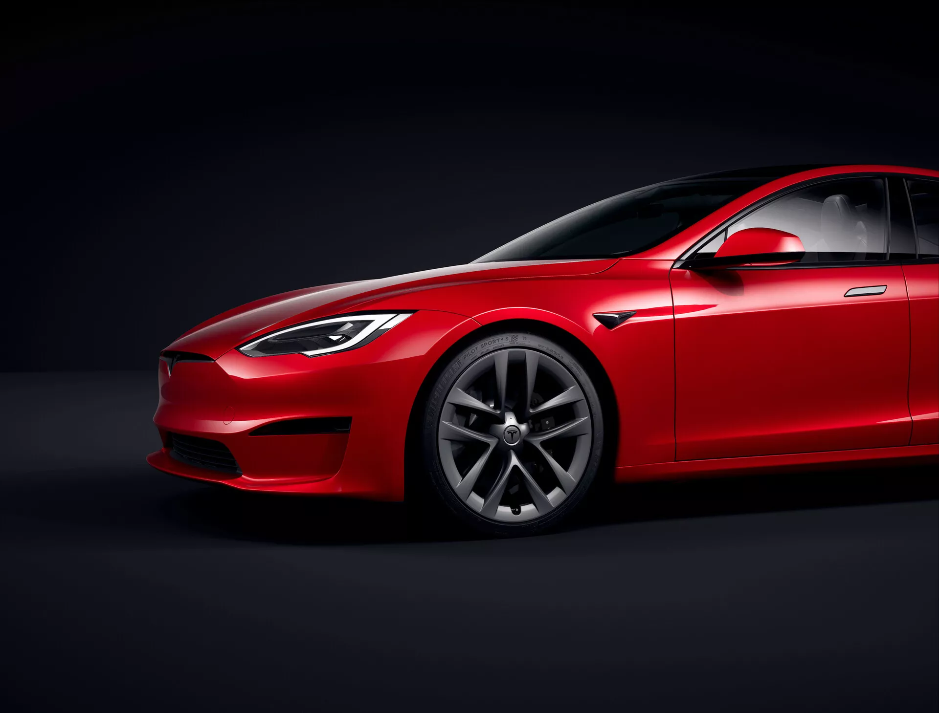 Tesla Model S 2016 (restylage 1ère génération) berline rouge en studio avec éclairage contrôlé et fond noir, vue en angle bas accentuant le profil aérodynamique et la garde au sol, vue de trois quarts avant montrant le côté conducteur, les phares, la calandre, une partie du capot, le design des jantes en alliage premium à motif multirayons, le pare-brise avec vitrage teinté, la ligne de toit inclinée, le rétroviseur, les poignées de porte et la ligne de fenêtres s'intégrant harmonieusement dans le design élégant, soulignant la finition de la peinture, les lignes de contour et les panneaux sculptés suggérant luxe et performance, éclairage focalisé mettant en valeur les détails du design.