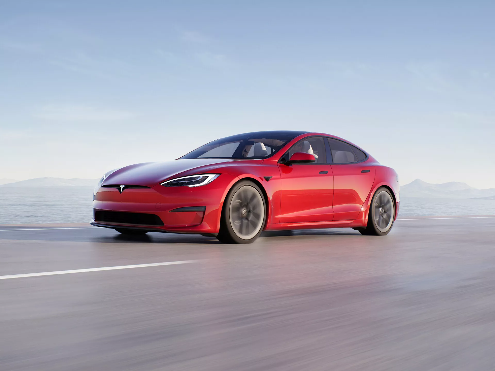 L'image montre une berline moderne à quatre portes Tesla Model S 2016 (facelift de la première génération) en mouvement sur une route ou une piste lisse. Présentée d'une vue de profil latéral à angle bas, les lignes élégantes et aérodynamiques du véhicule sont clairement visibles, mettant en valeur sa calandre fermée distinctive, ses phares LED minces et son pare-chocs avant lisse. Le design fastback avec une ligne de toit doucement inclinée, des poignées de portes encastrées rétractables et des rétroviseurs latéraux aérodynamiques minimalistes mettent en avant l'accent mis sur l'aérodynamisme du véhicule. Les grandes roues à plusieurs branches suggèrent une optimisation du flux d'air et une réduction de la traînée. La composition utilise le flou de mouvement pour transmettre une haute vitesse, avec la voiture elle-même nettement mise au point contre un arrière-plan flou. Cette photographie professionnelle souligne la nature électrique de la voiture, son éclairage avancé, son design aérodynamique et ses caractéristiques potentielles d'assistance au conducteur.