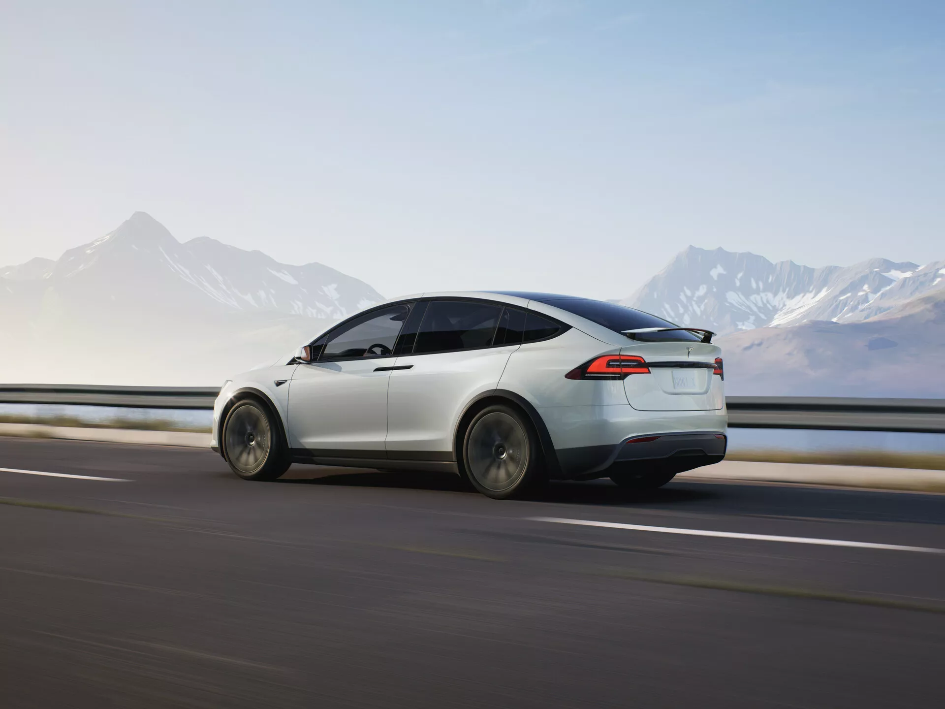 Vue dynamique arrière trois quarts d'une Tesla Model X 2021 (1ère génération-restylée) en mouvement, mettant en évidence son design aérodynamique, sa peinture blanche élégante, ses feux arrière à LED et son design moderne des roues. La voiture est capturée en mouvement de gauche à droite contre une route floue et une glissière de sécurité, suggérant la vitesse, avec un horizon montagneux en arrière-plan. L'éclairage doux indique tôt le matin ou en fin d'après-midi, mettant en valeur les lignes électriques épurées et les fonctionnalités avancées du véhicule dans un cadre pittoresque.